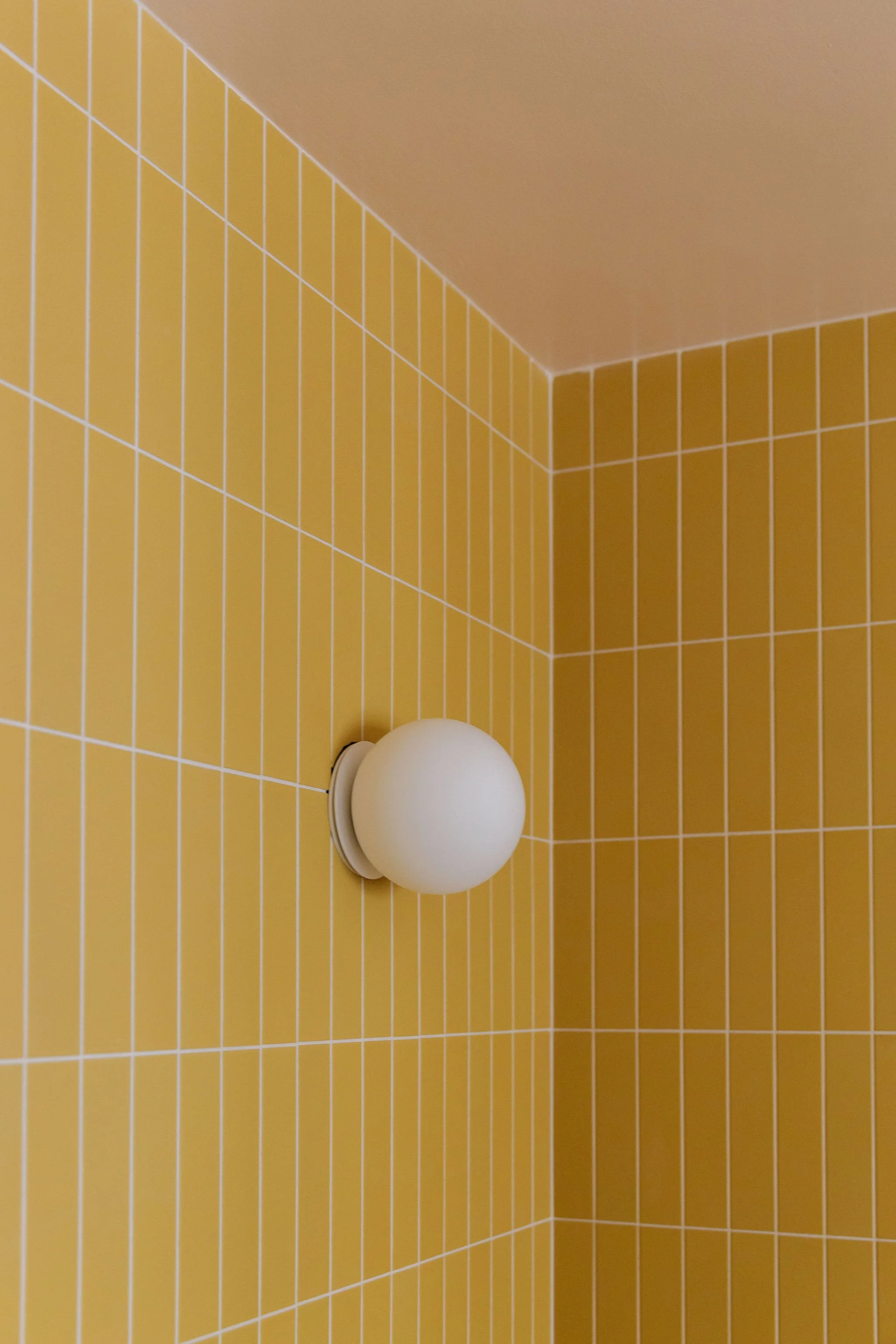 A white spherical sconce mounted on a yellow tiled bathroom wall.