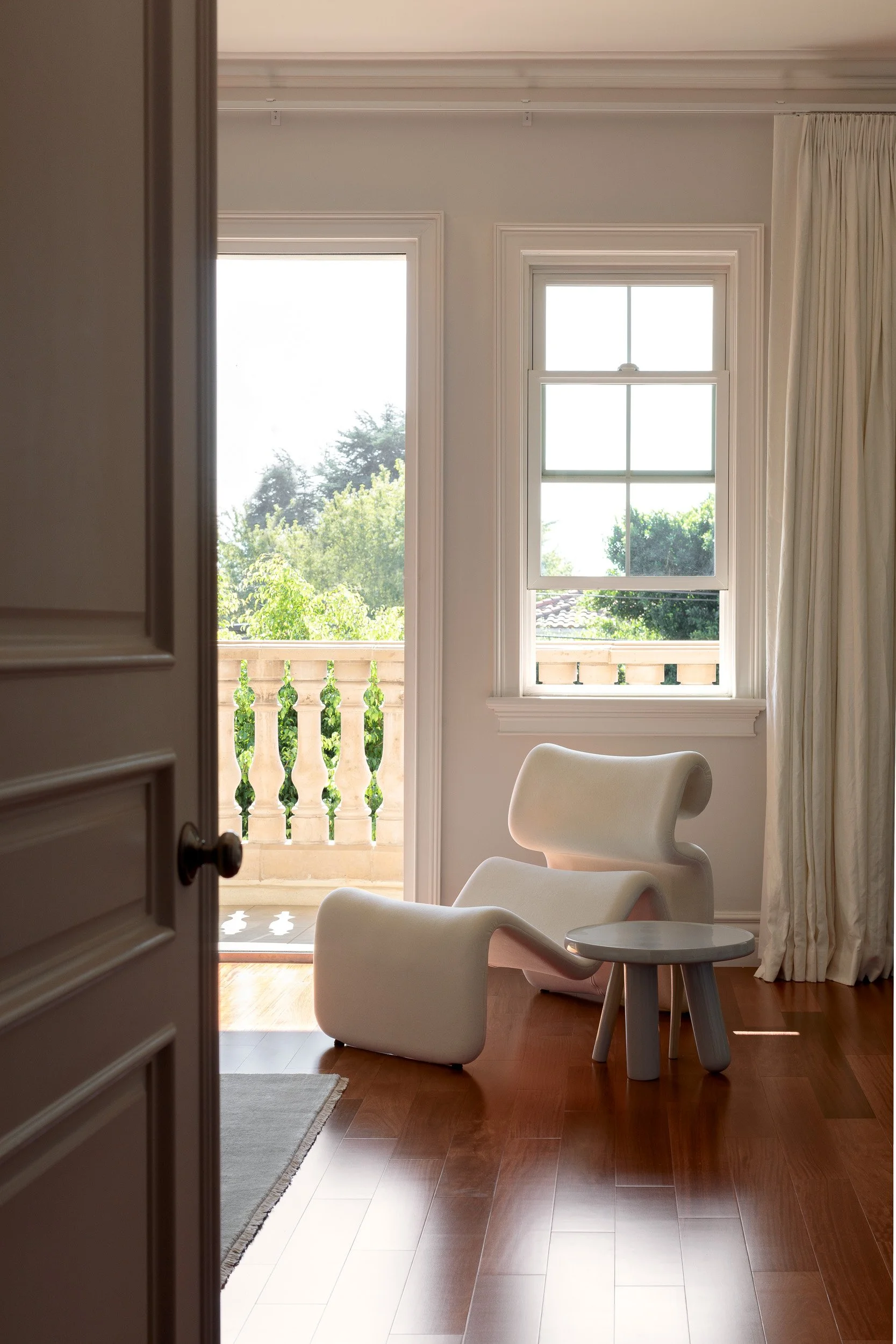 Interior of a room viewed through an open door, featuring a modern white chair, a small round table, hardwood floors, a window with curtains, and a balcony with a decorative stone railing outside.