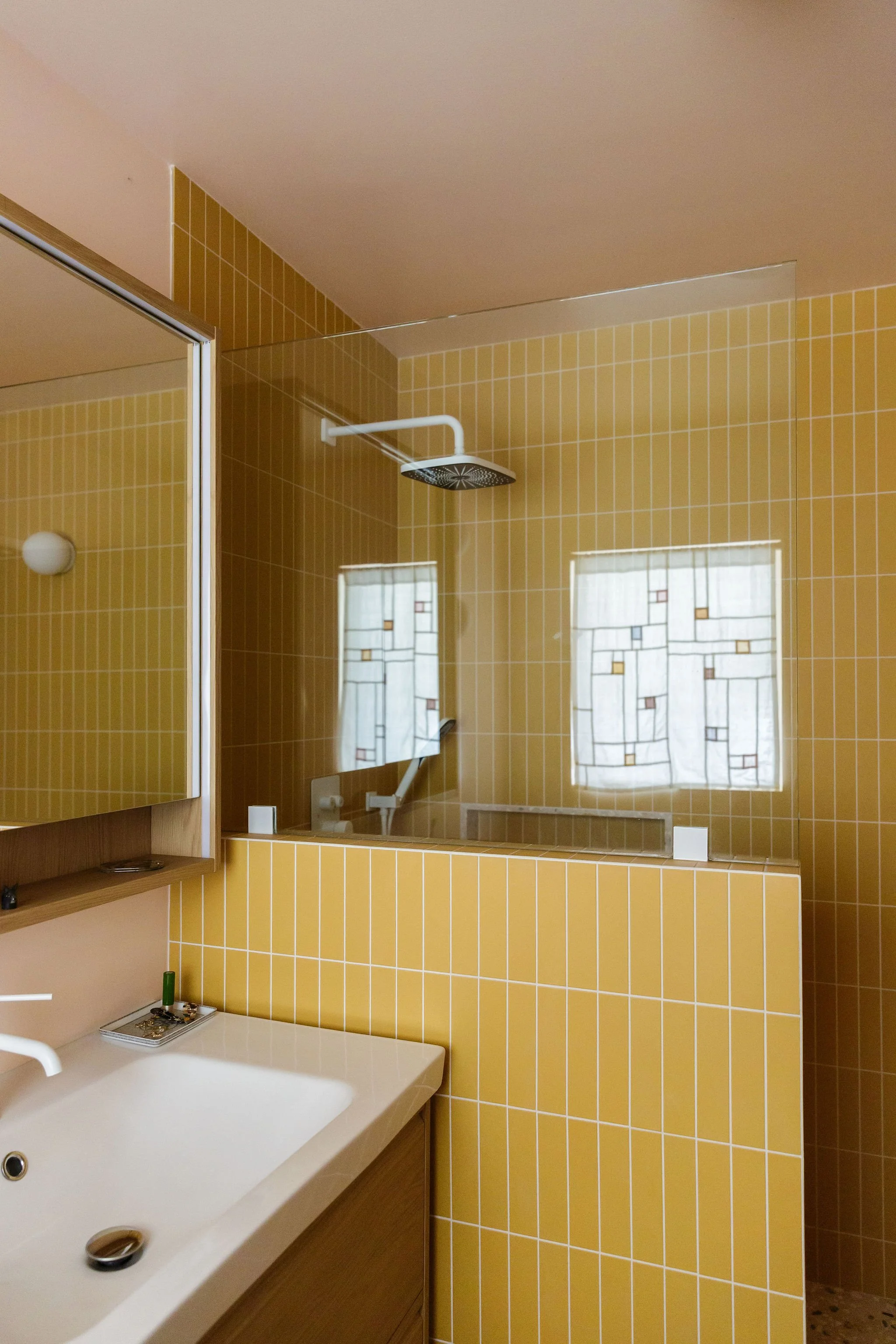 A bathroom with yellow tiled walls, a mirror, a showerhead, a white sink, and a window with patterned curtains.