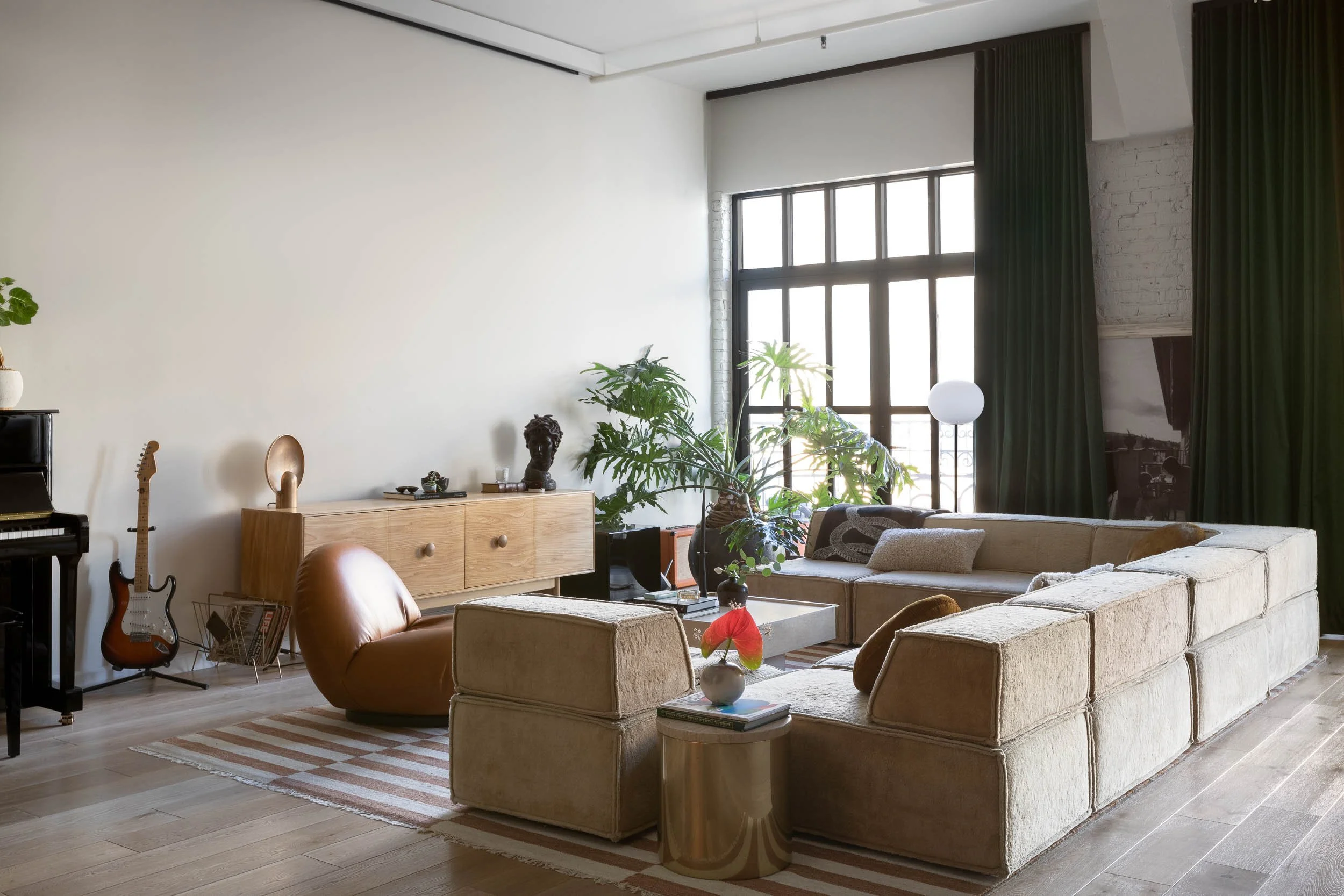 Living room with a beige sectional sofa, a brown chair, a striped rug, a wooden sideboard, a guitar, and green plants near large windows with dark curtains.