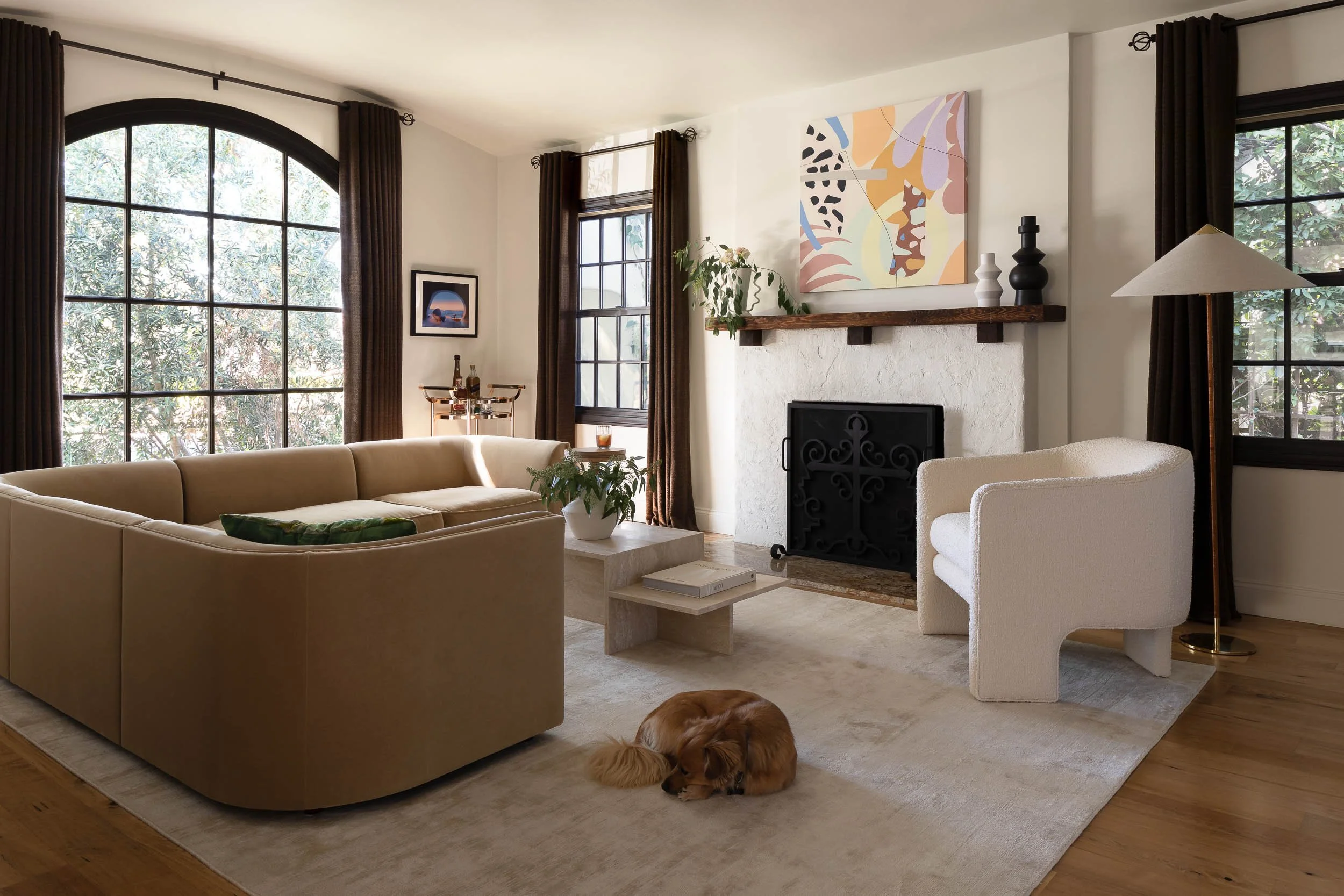 Living room with large arched and rectangular windows, beige sofa, white armchair, fireplace with painting above, potted plant, artwork, and a dog lying on the carpet.