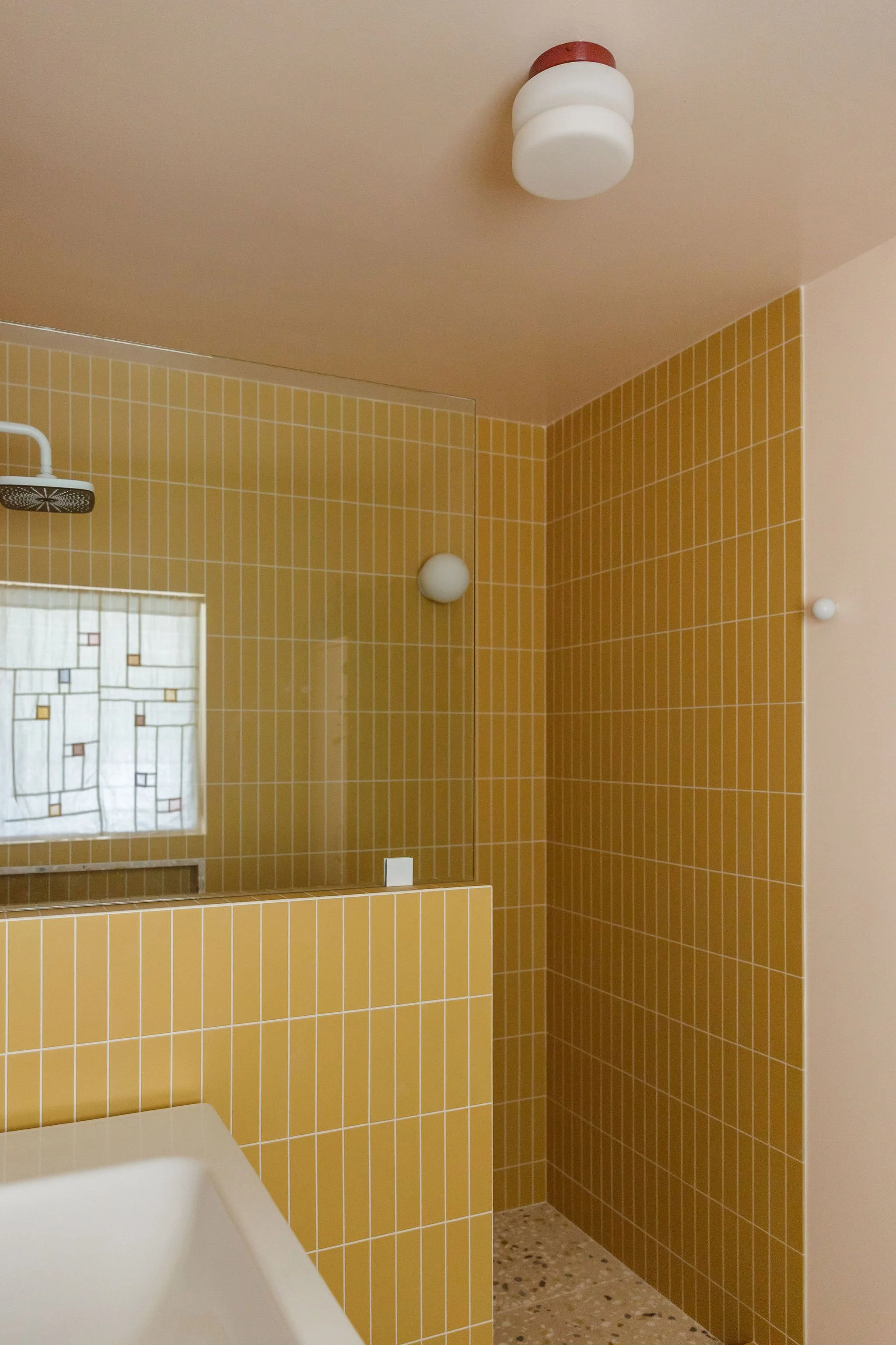 A bathroom with yellow square tiles on the wall and a a large mirror covering the entire wall. A showerhead is mounted on the wall. The bathroom has a pale pink terrazzo floor and two round white wall lights, with one on the ceiling.