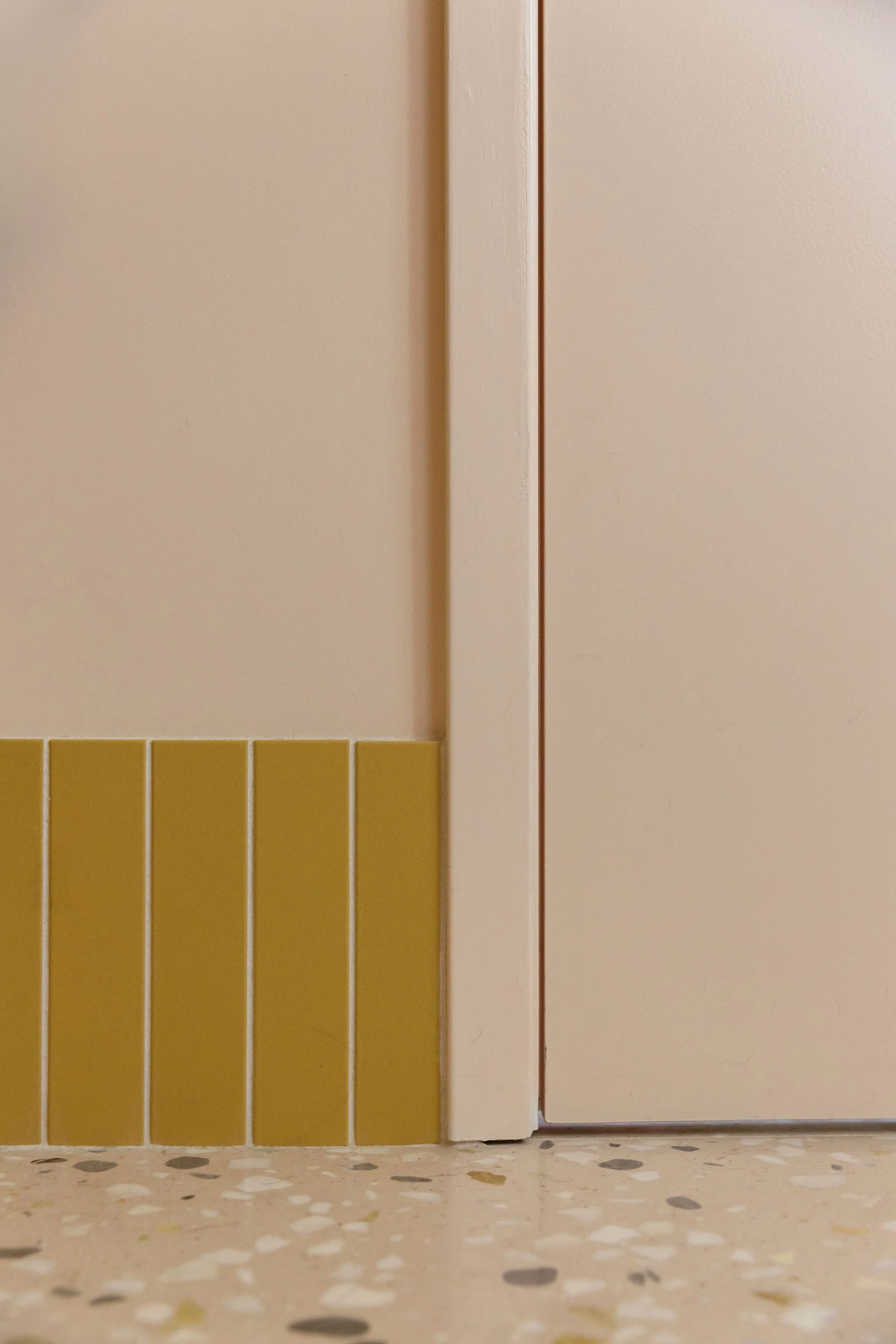 Close-up of a yellow tiled wall with vertical pink door and a terrazzo floor.