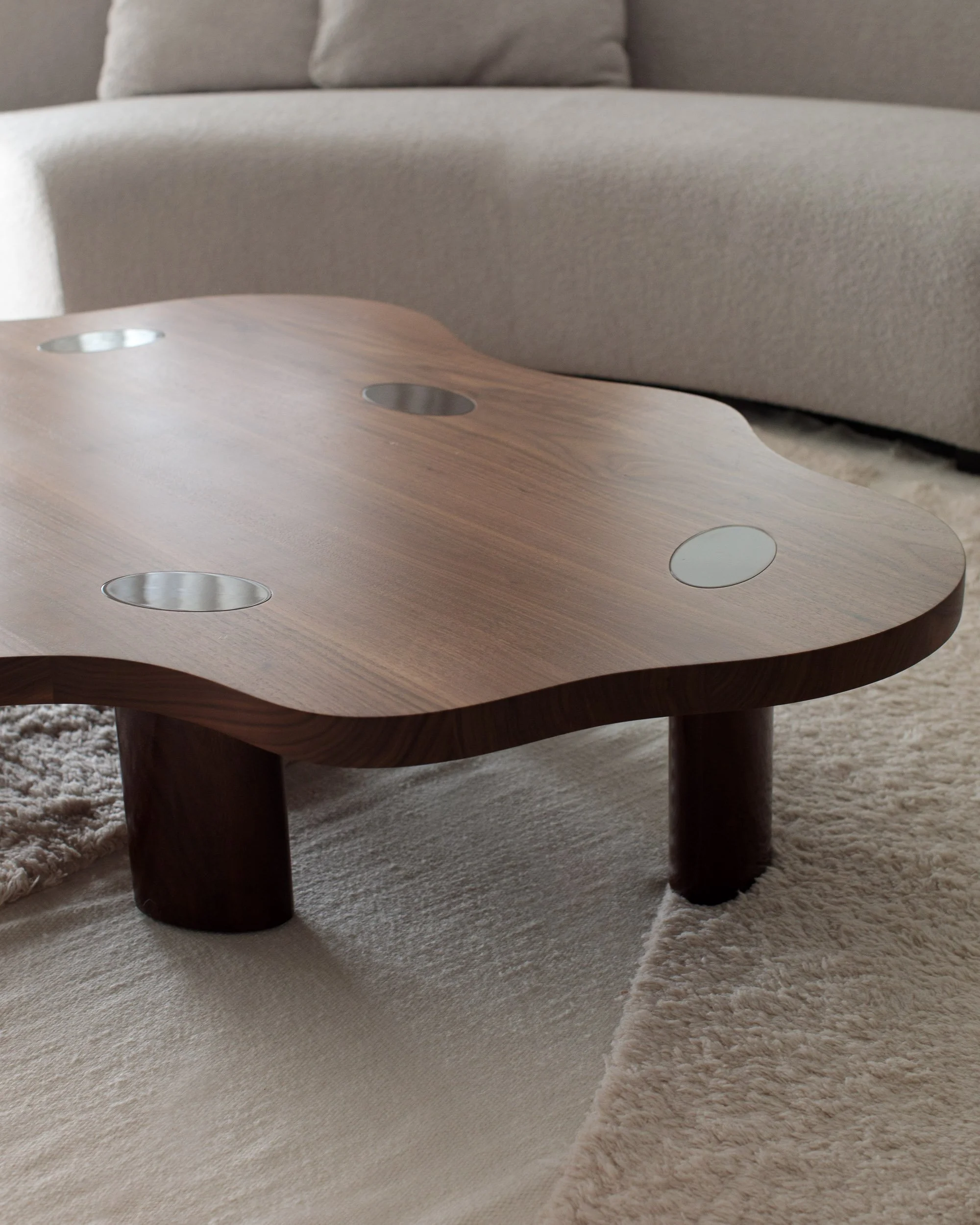 A modern wooden coffee table with circular metal insets, situated on a beige shaggy rug in front of a beige sofa.