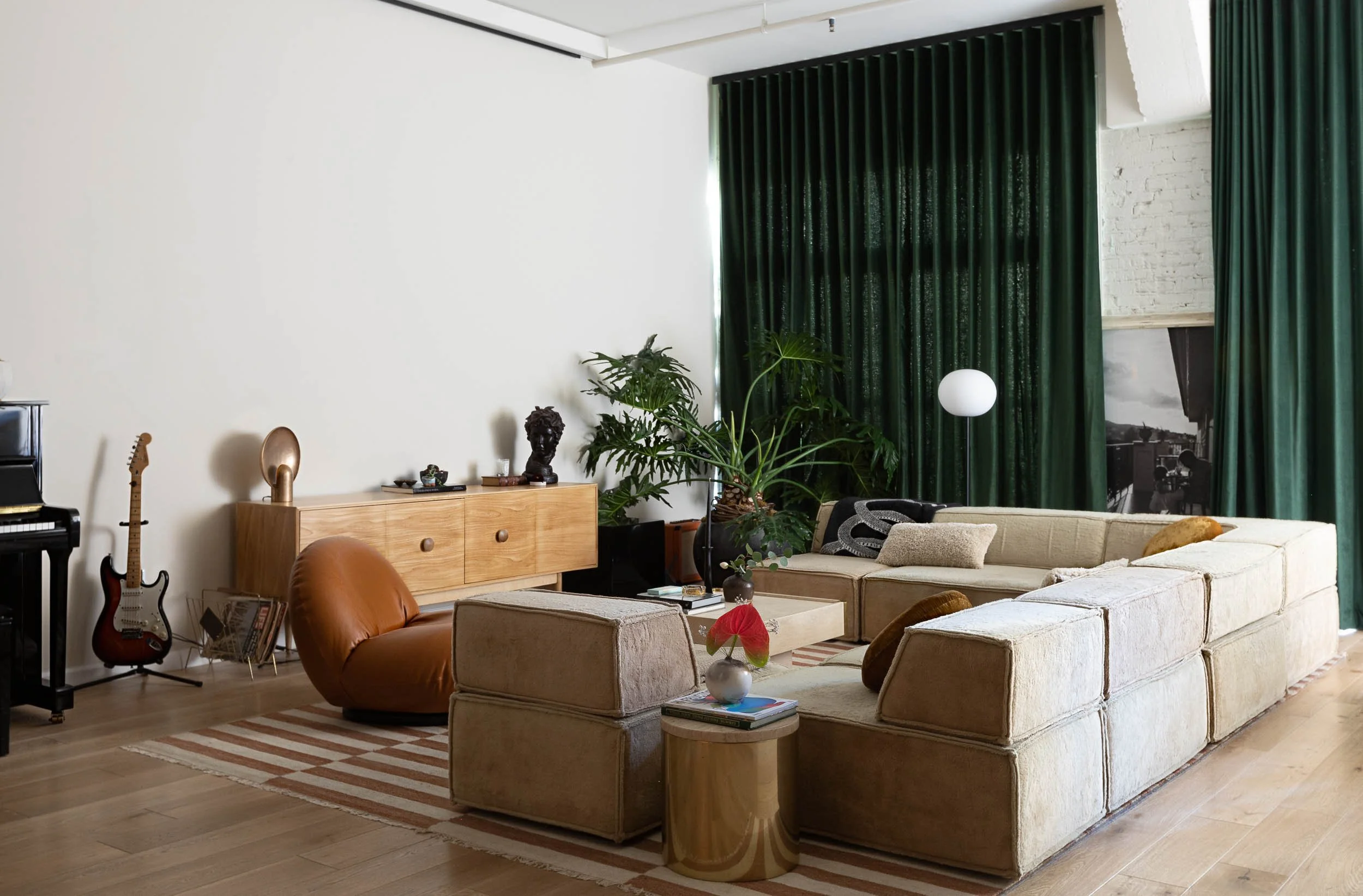 Living room with beige sectional sofa, green curtains, potted plants, guitar, and wooden furniture.