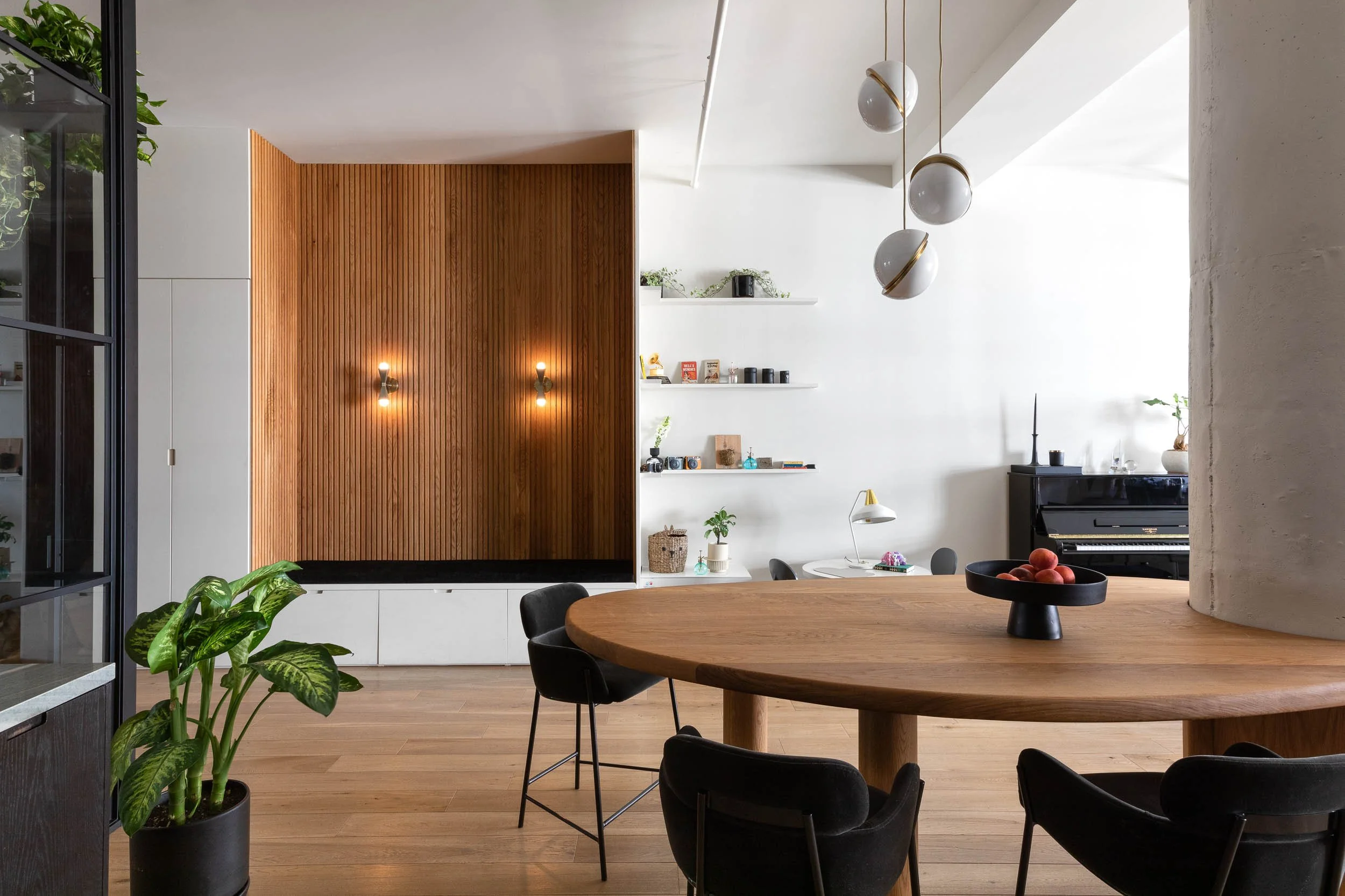 Modern dining room with a round wooden table, black chairs, a black piano, white walls with floating shelves, a wooden accent wall, and a potted plant.