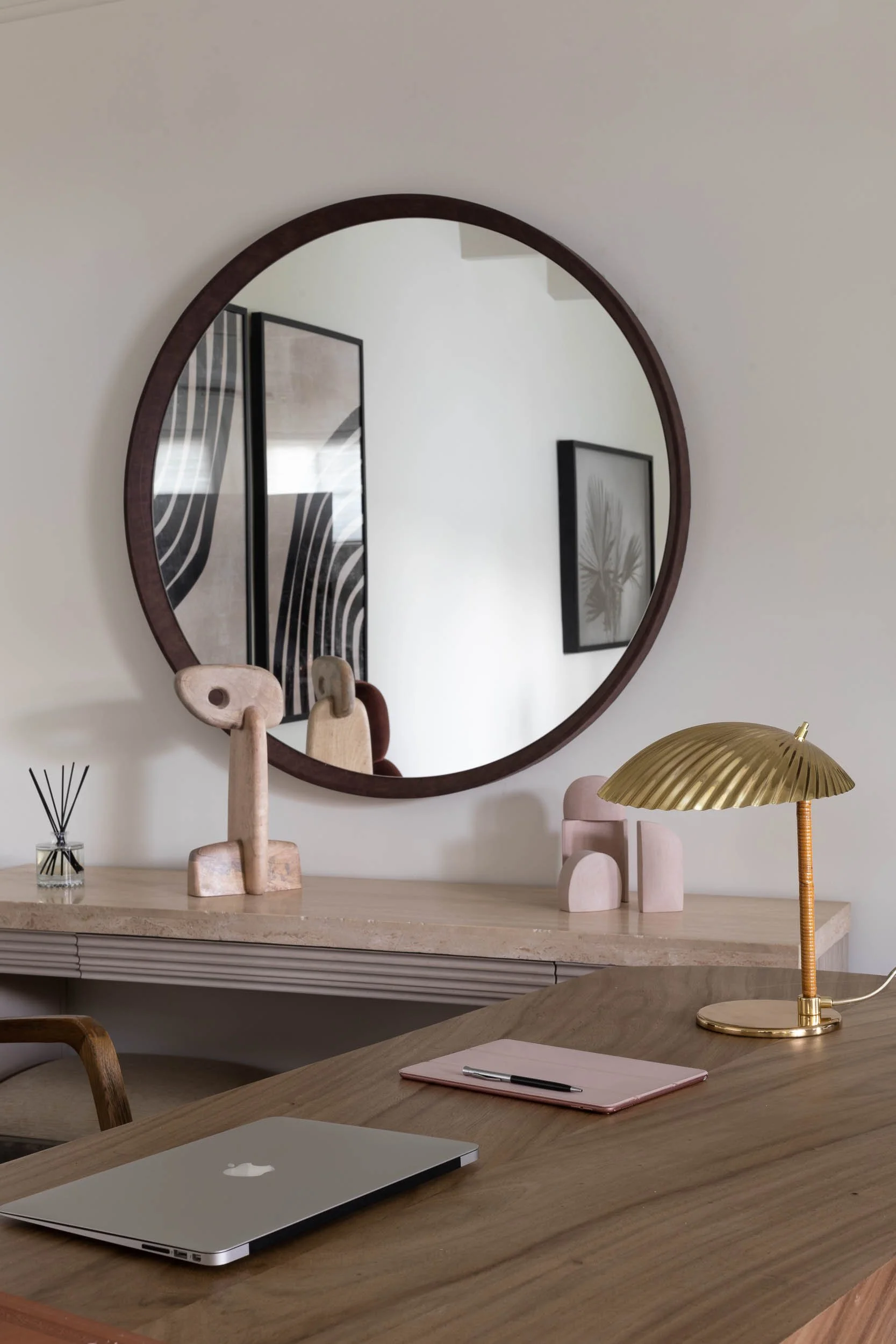 Wooden desk with closed silver MacBook, pink notebook, and silver pen, with a gold table lamp, in front of a large round mirror reflecting modern wall art and decor.