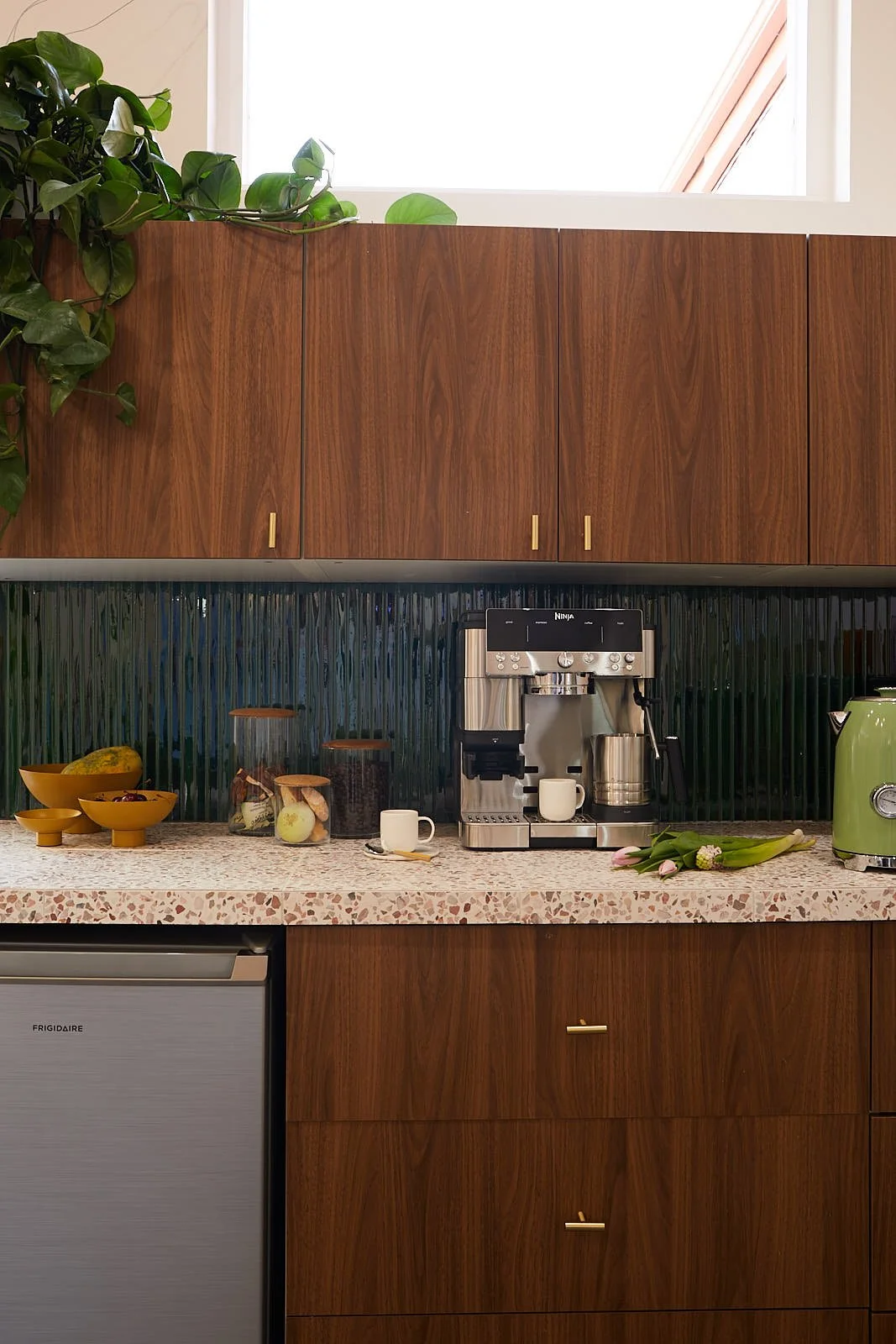Kitchen counter with a coffee machine, a small cup, and various items such as bowls of fruit, jars, a tulip bouquet, and a green toaster.