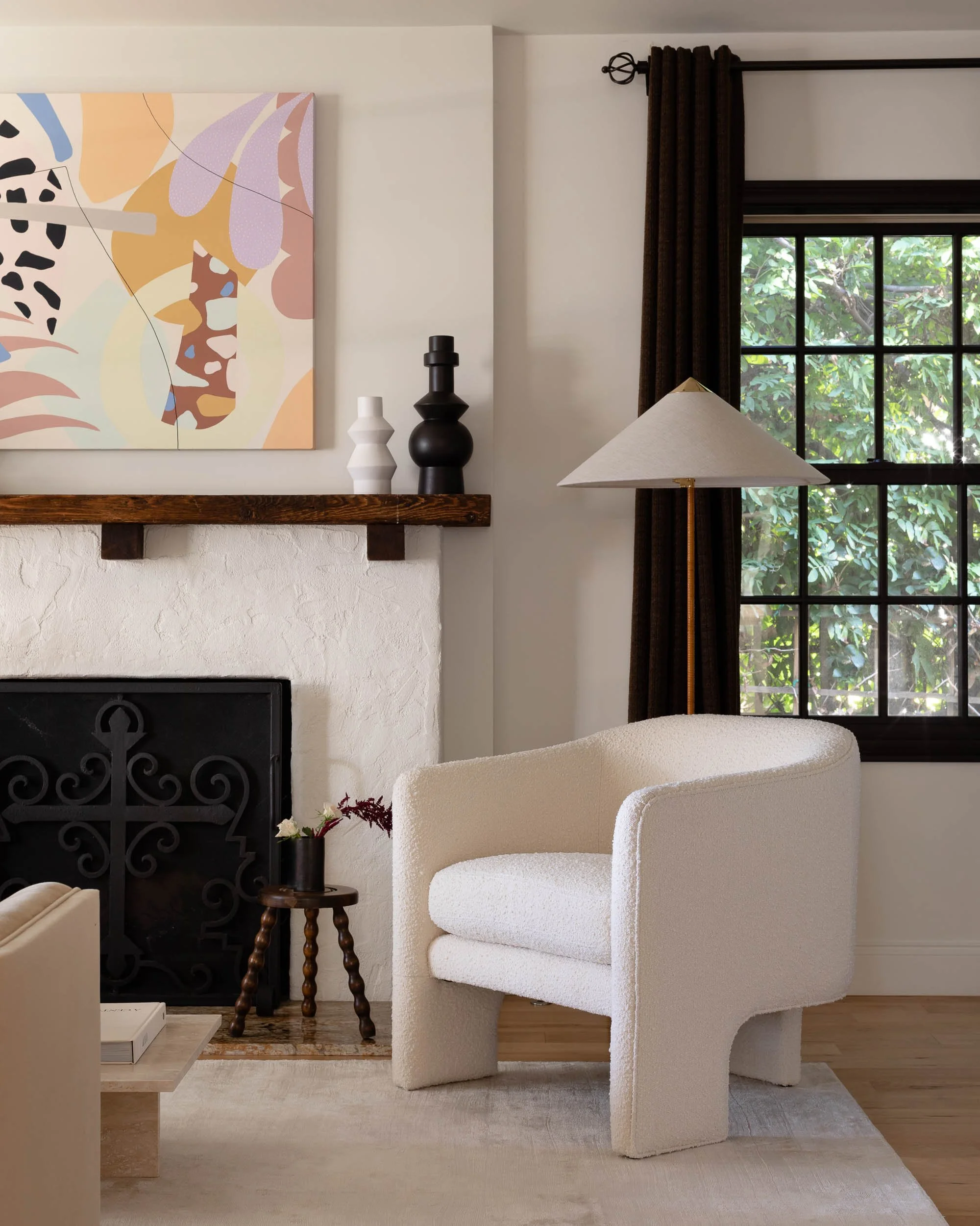 A cozy living room corner featuring a white textured armchair, a small wooden side table with flowers, a fireplace with decorative black iron grate, a window with dark brown curtains, a standing lamp with a beige shade, a colorful abstract painting a