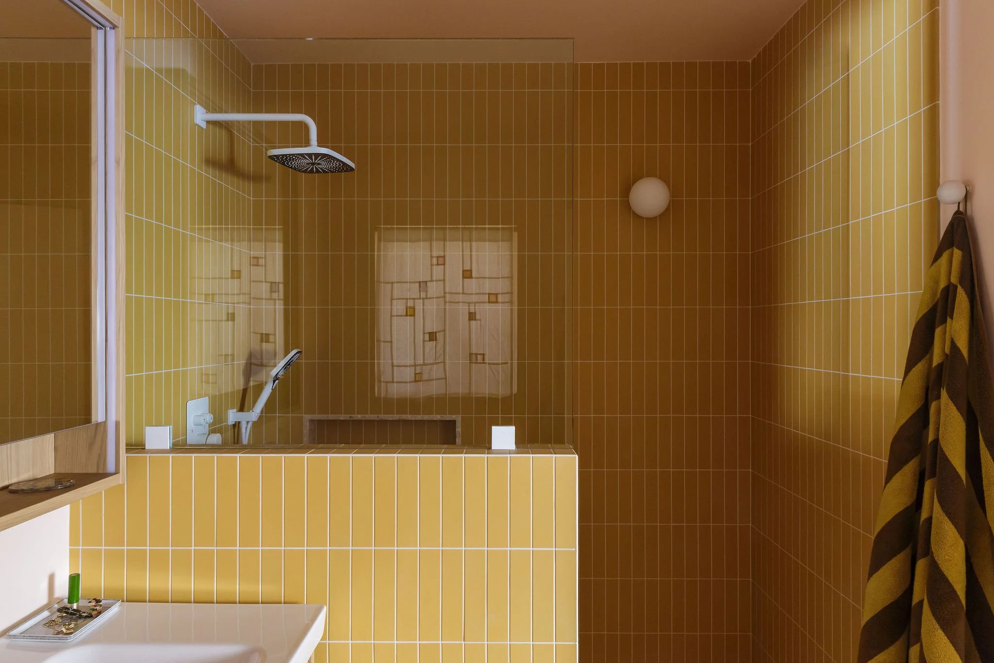 Shower area with yellow tiles, a white shower head, and a glass partition. There is a window with curtains, a towel hanging, and a small shelf with toiletries.