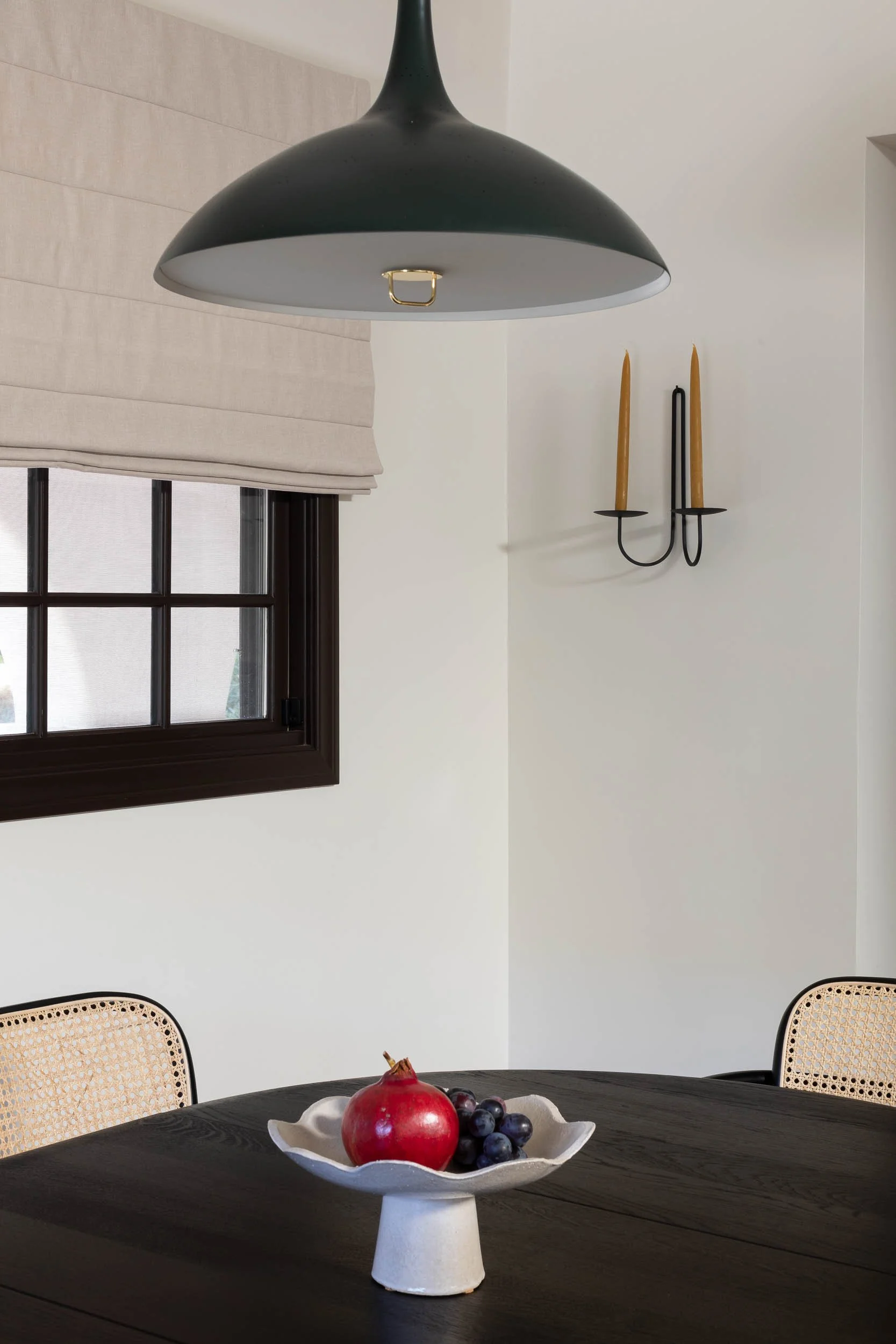A dining room with a dark wooden table, beige woven chairs, a white ceramic fruit bowl with grapes and a pomegranate, a window with a beige Roman shade, and a black pendant light fixture hanging from the ceiling.