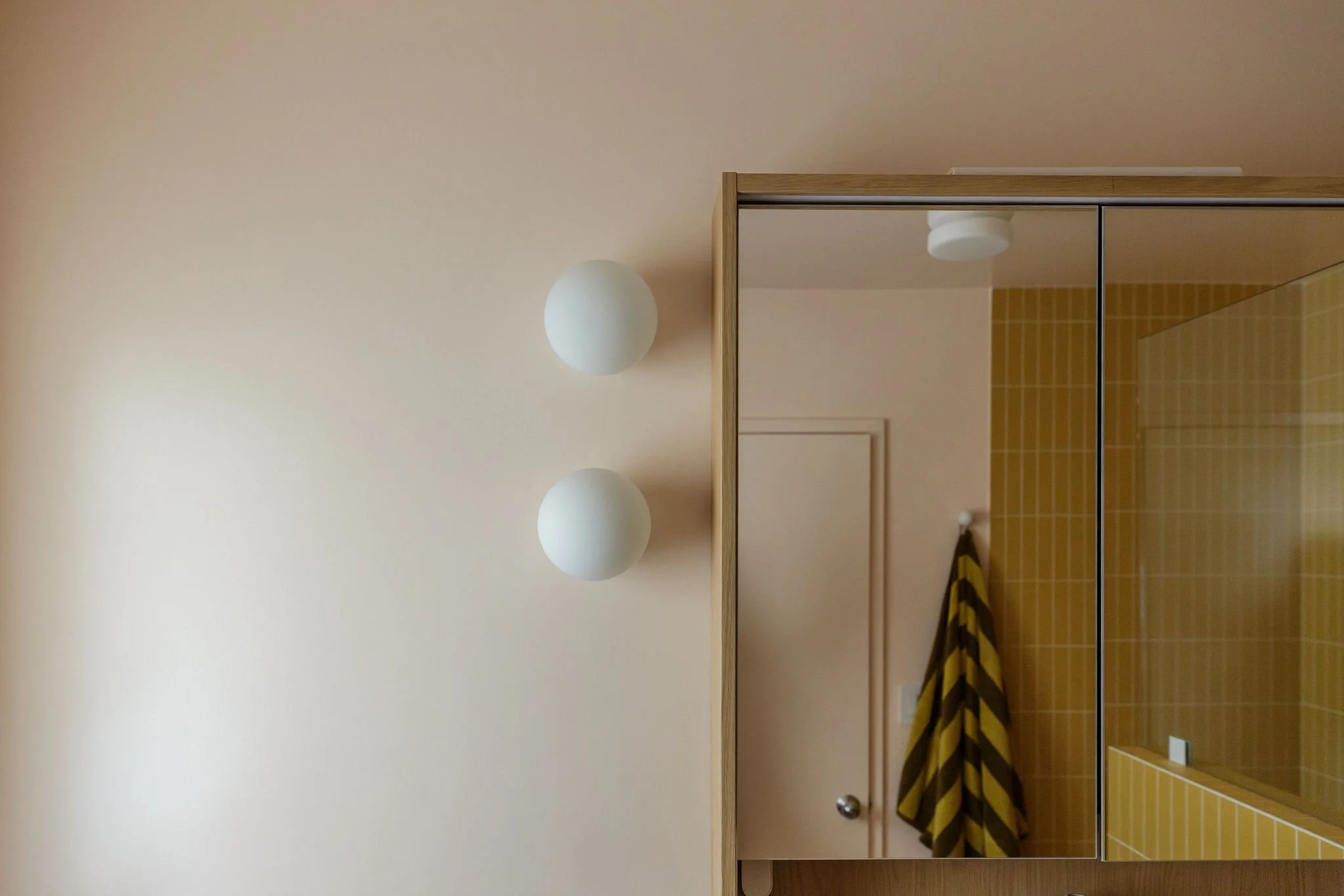 View of a bathroom with a large mirror, yellow tile walls, a partial view of a towel with yellow and brown stripes, and a pink wall with two white round light fixtures.