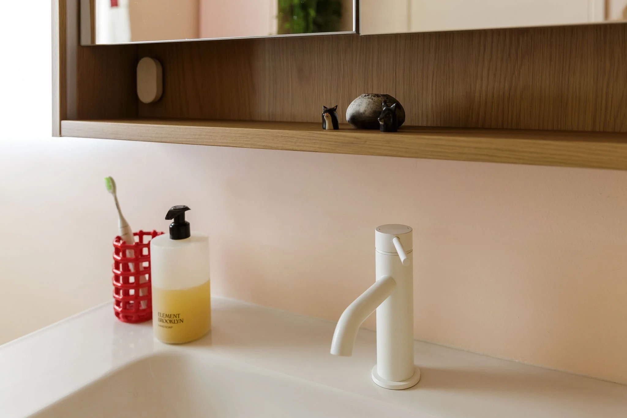 Bathroom sink with a white faucet, yellow soap dispenser, and a red toothbrush holder on a white countertop. A wooden shelf with decorative animal figurines is visible above the sink.