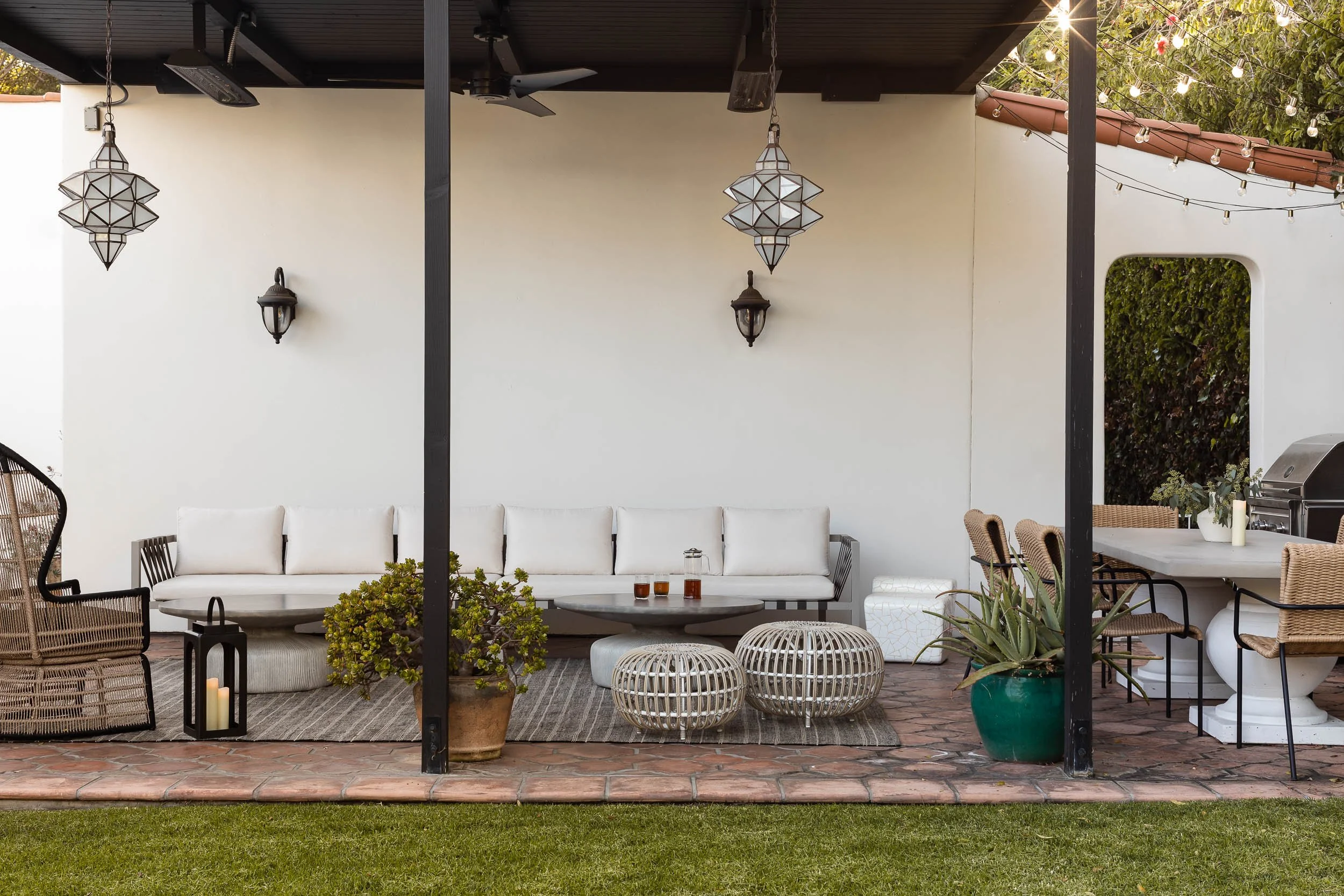 Outdoor patio with white cushioned sofa, round woven coffee tables, wicker chairs, potted plants, hanging lanterns, and string lights under a dark wooden ceiling.