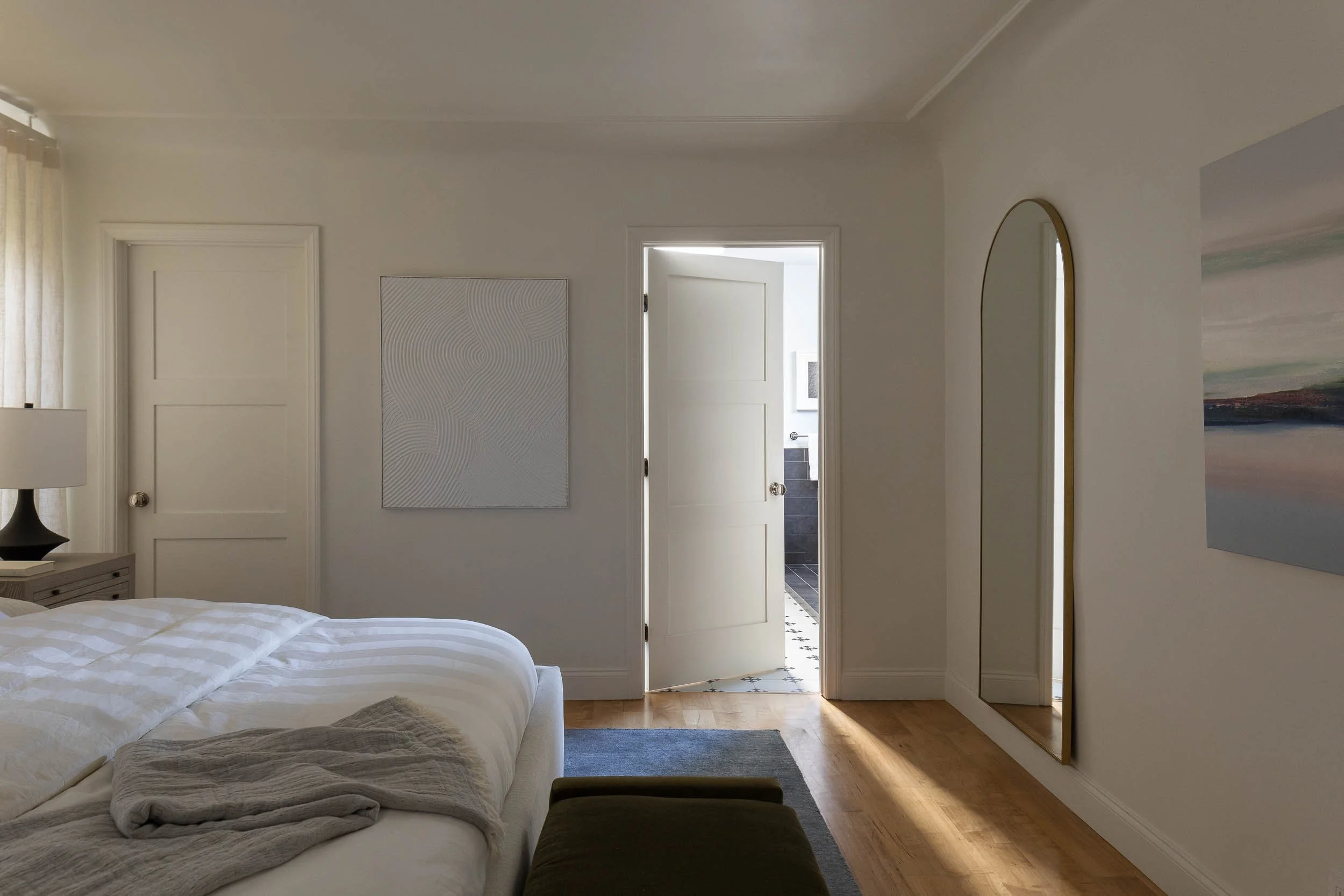 A bedroom with a partially visible bed, a bedside table with a lamp, a mirror on the wall, and an open door leading to a bathroom.