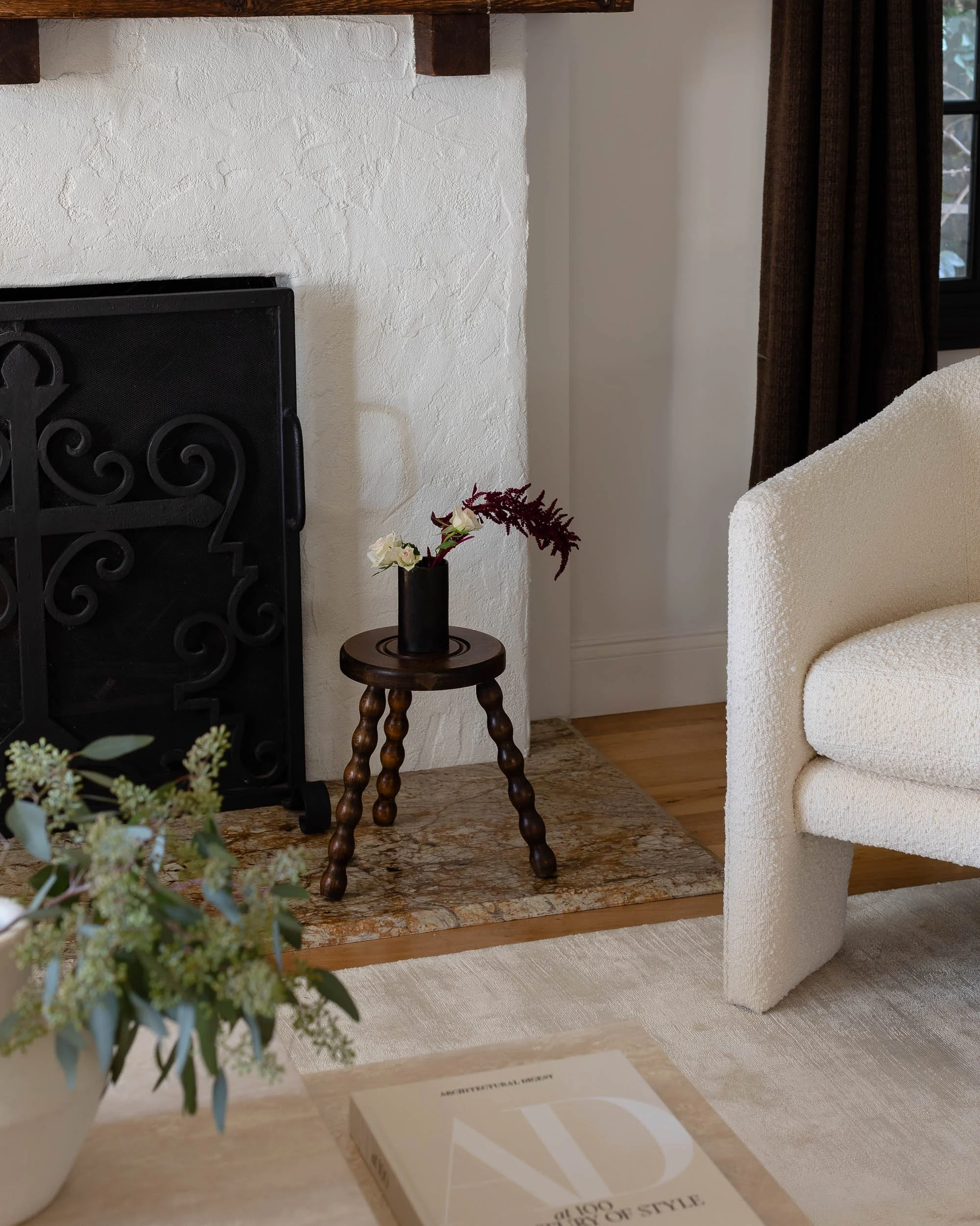 A cozy living room corner featuring a black fireplace with a decorative wrought iron screen, a small wooden side table with a black vase holding white and red flowers, a white armchair with textured fabric, a dark curtain, a book on a light-colored r