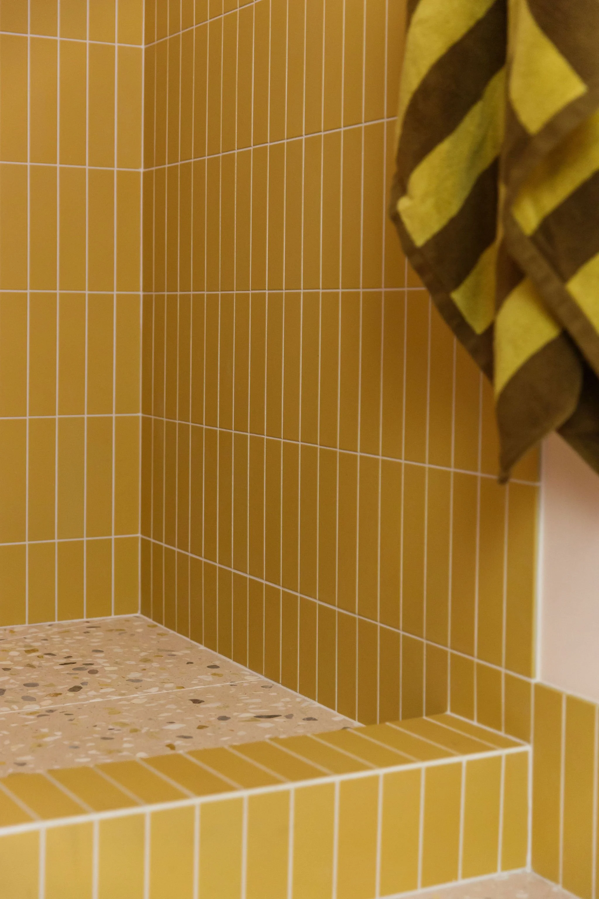 Interior view of a shower with yellow tiles on the walls and a terrazzo floor. A brown and yellow striped towel hangs on the right side.