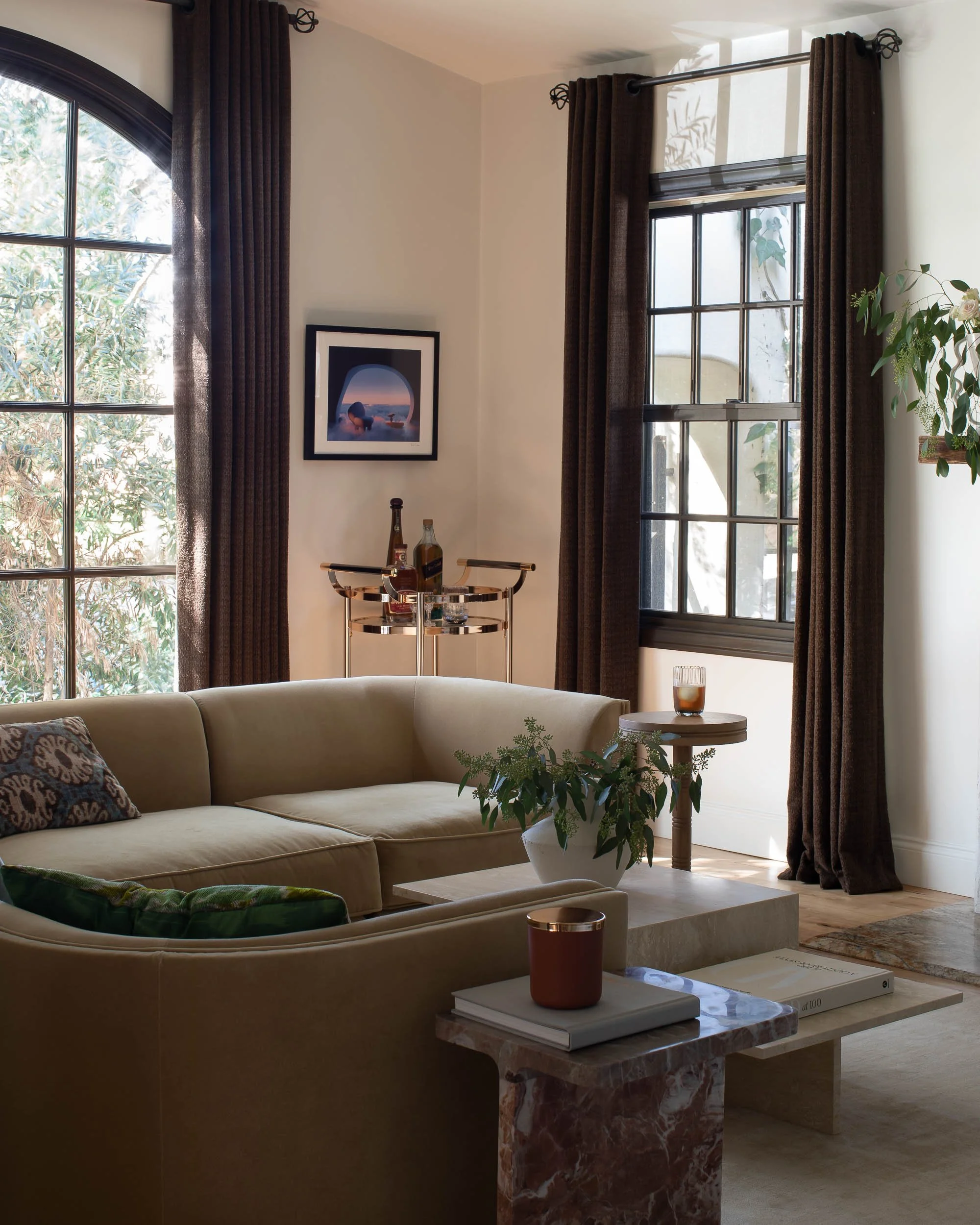 Living room with beige sofa, green and patterned pillows, marble and wood coffee tables, plants, large windows with brown curtains, framed artwork, and a bar cart with bottles in the corner.