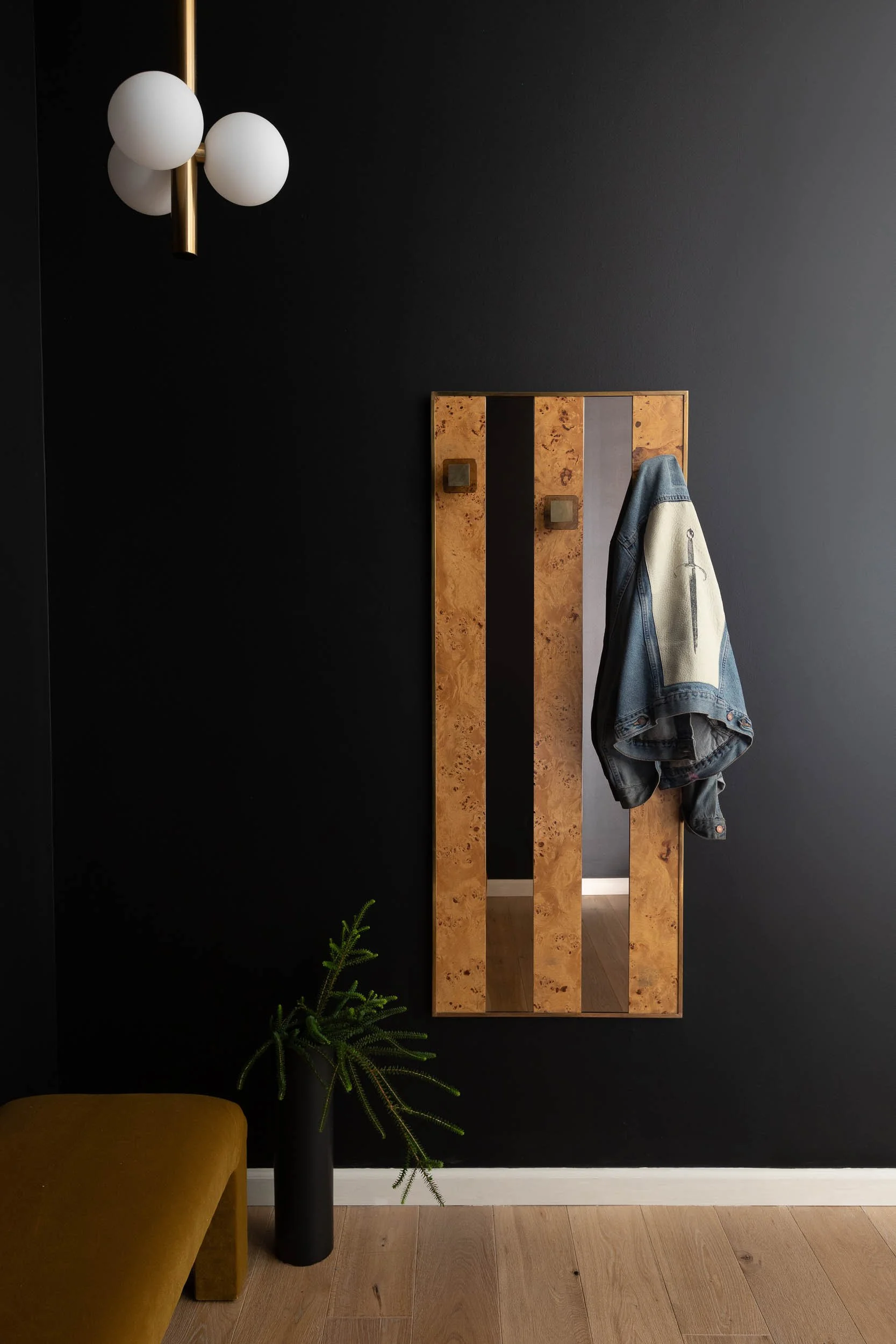 A wall with black paint, a wooden mirror with hooks, a denim jacket hanging, a modern wall light fixture with three white globes and brass accents, a small potted plant in a black vase, and part of a mustard-colored chair.