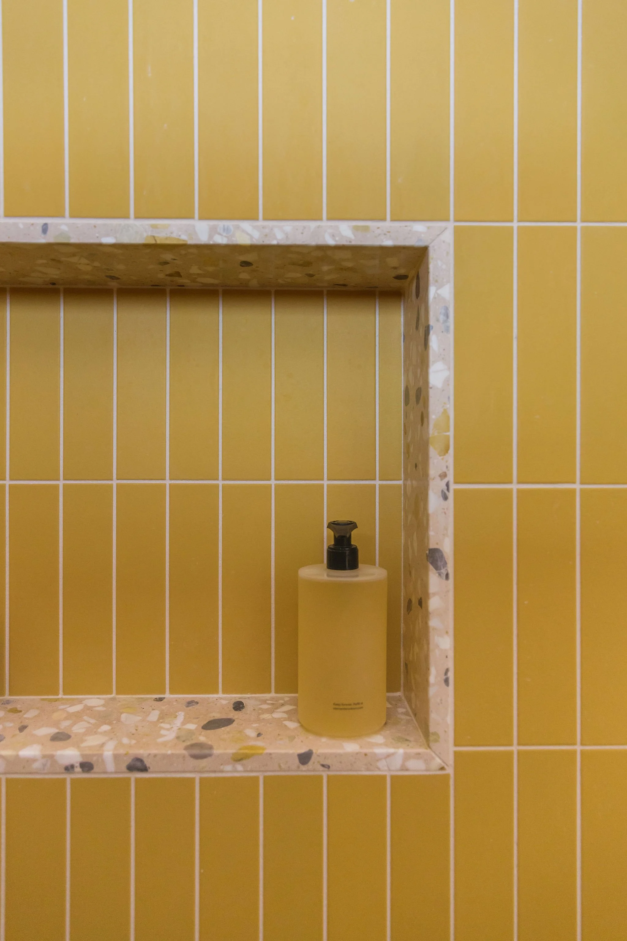 Bathroom shelf with a yellow bottle, yellow soap dispenser, and yellow wall tiles with white grout lines.