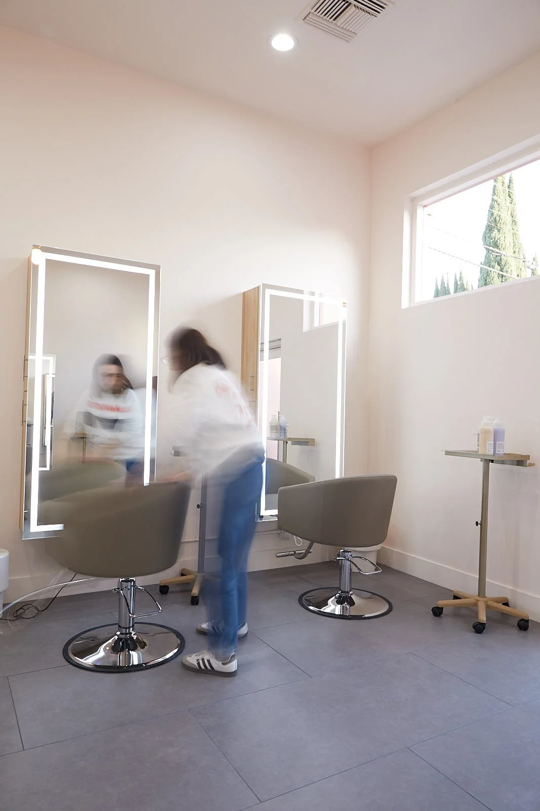 A person in a salon or beauty studio standing in front of a mirror with LED lighting, next to a salon chair and a small rolling table with hair or beauty products. The background shows a large window and ceiling vent.