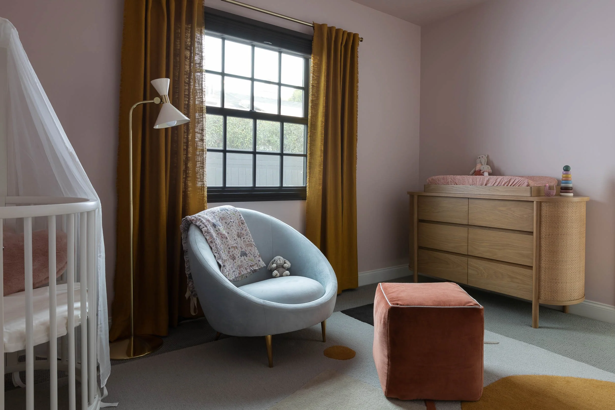A cozy nursery with a white crib, a light blue rocking chair with a blanket and stuffed animal, a pink ottoman, a wooden dresser with a pink changing pad and plush toy on top, and a window with mustard-colored curtains.