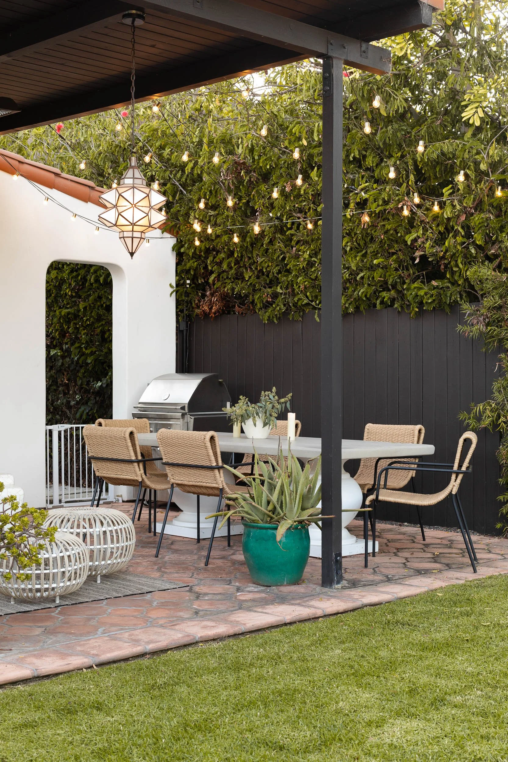 Outdoor patio with string lights and a decorative hanging lantern, beige wicker chairs around a white table, potted aloe vera plant in a green pot, brick tiled floor, lush green trees and black fencing in the background.