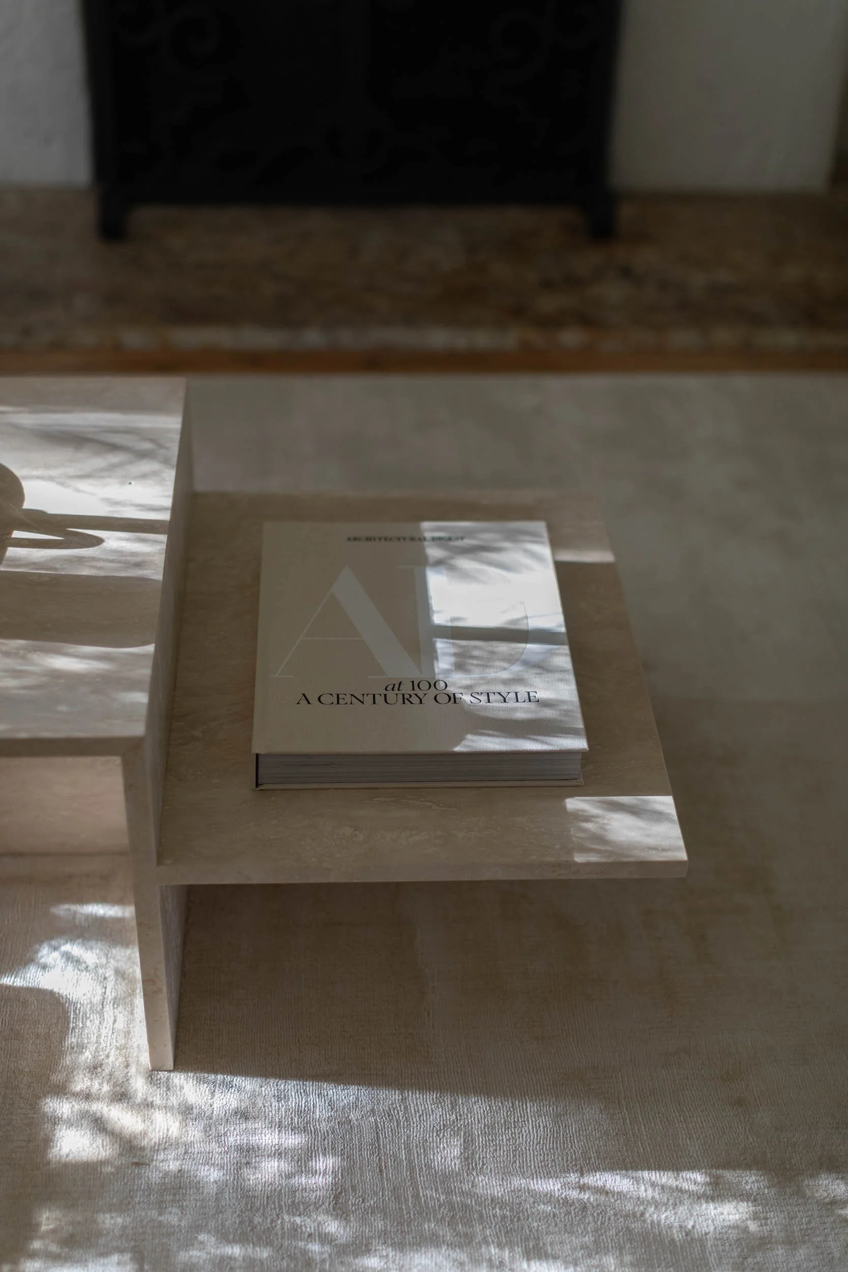 Book titled "At 100: A Century of Style" placed on a beige table with sunlight creating shadows.