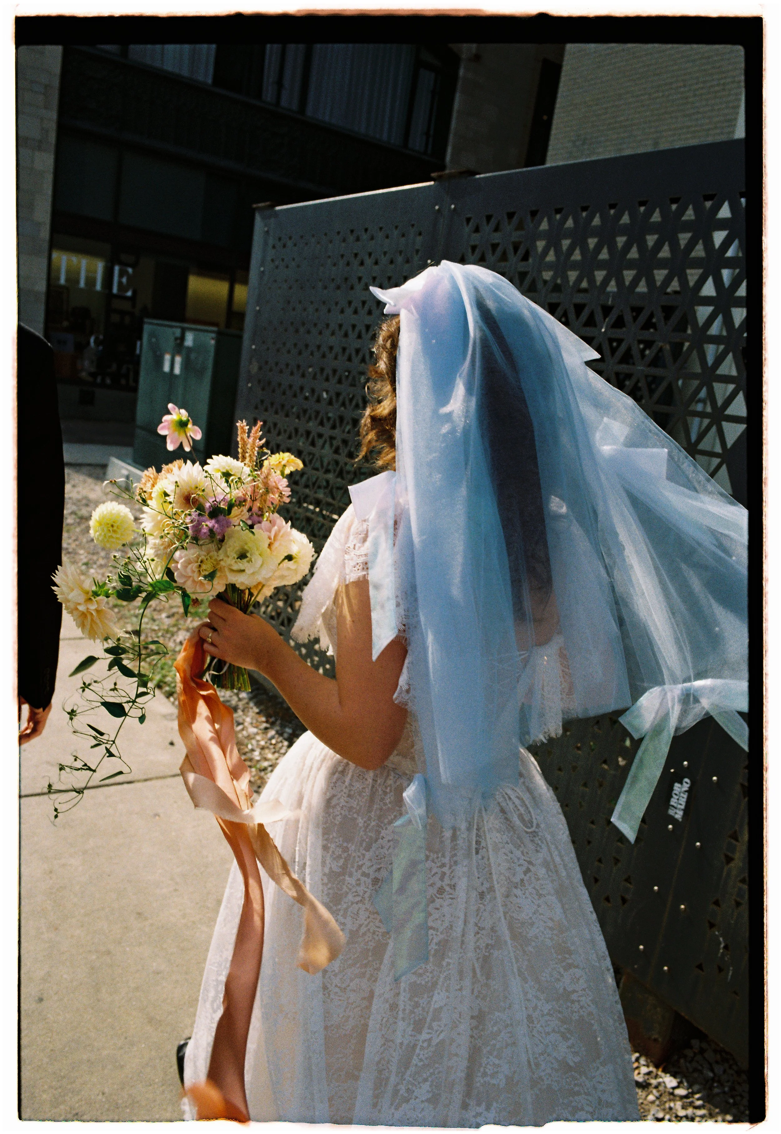 A bride in a white lace wedding dress with a long veil holding a bouquet of pink, white, and yellow flowers with ribbons, standing outdoors near a black lattice fence.