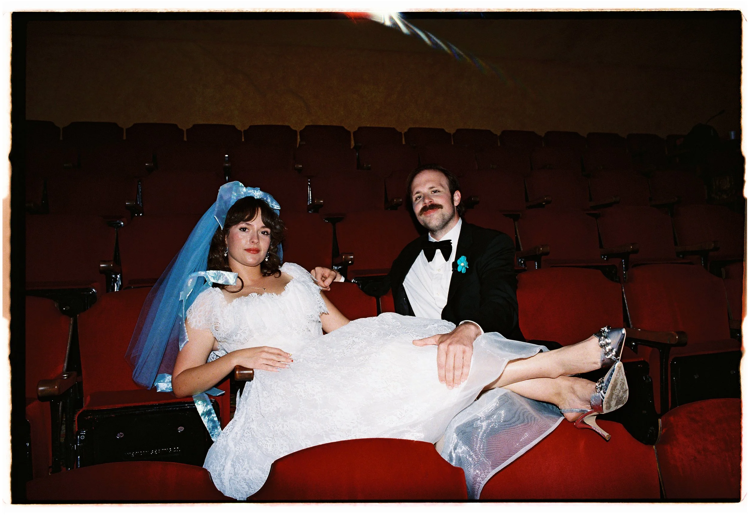 A woman in a wedding dress and a man in a tuxedo sitting in a theatre, the woman lying across the seats with her legs resting on the man's lap. They are both smiling, and the woman is wearing high heels.