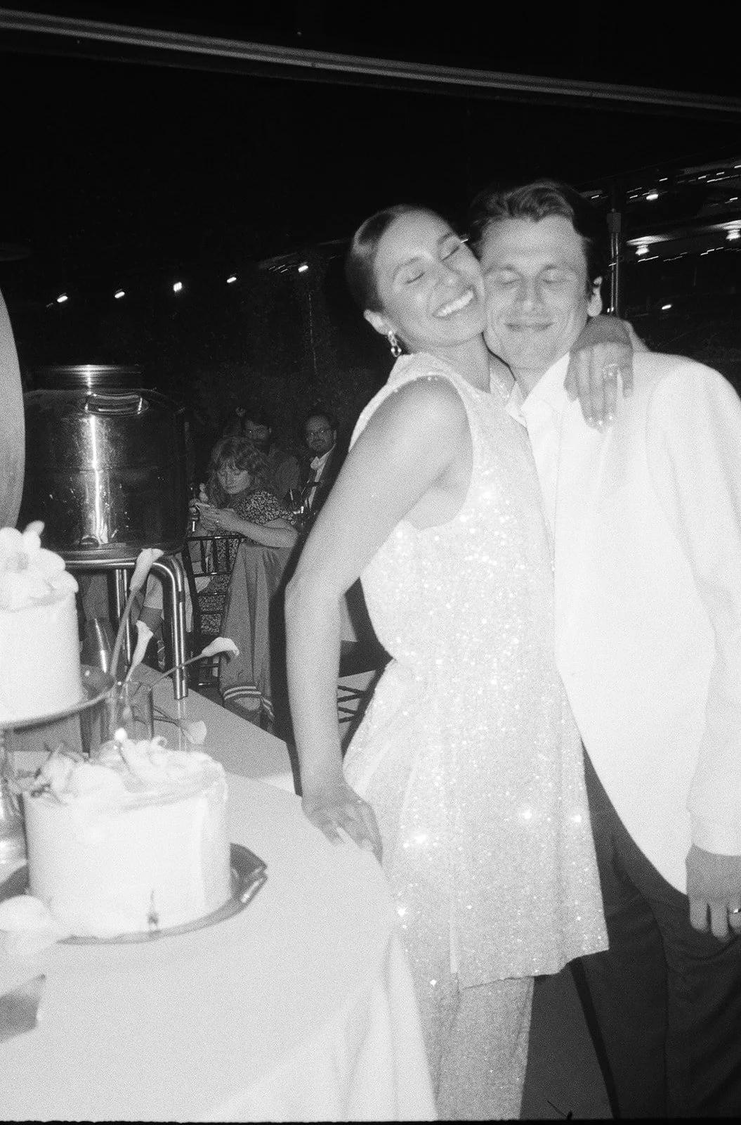 A woman and man smiling and hugging at a celebration with cake and desserts on the table