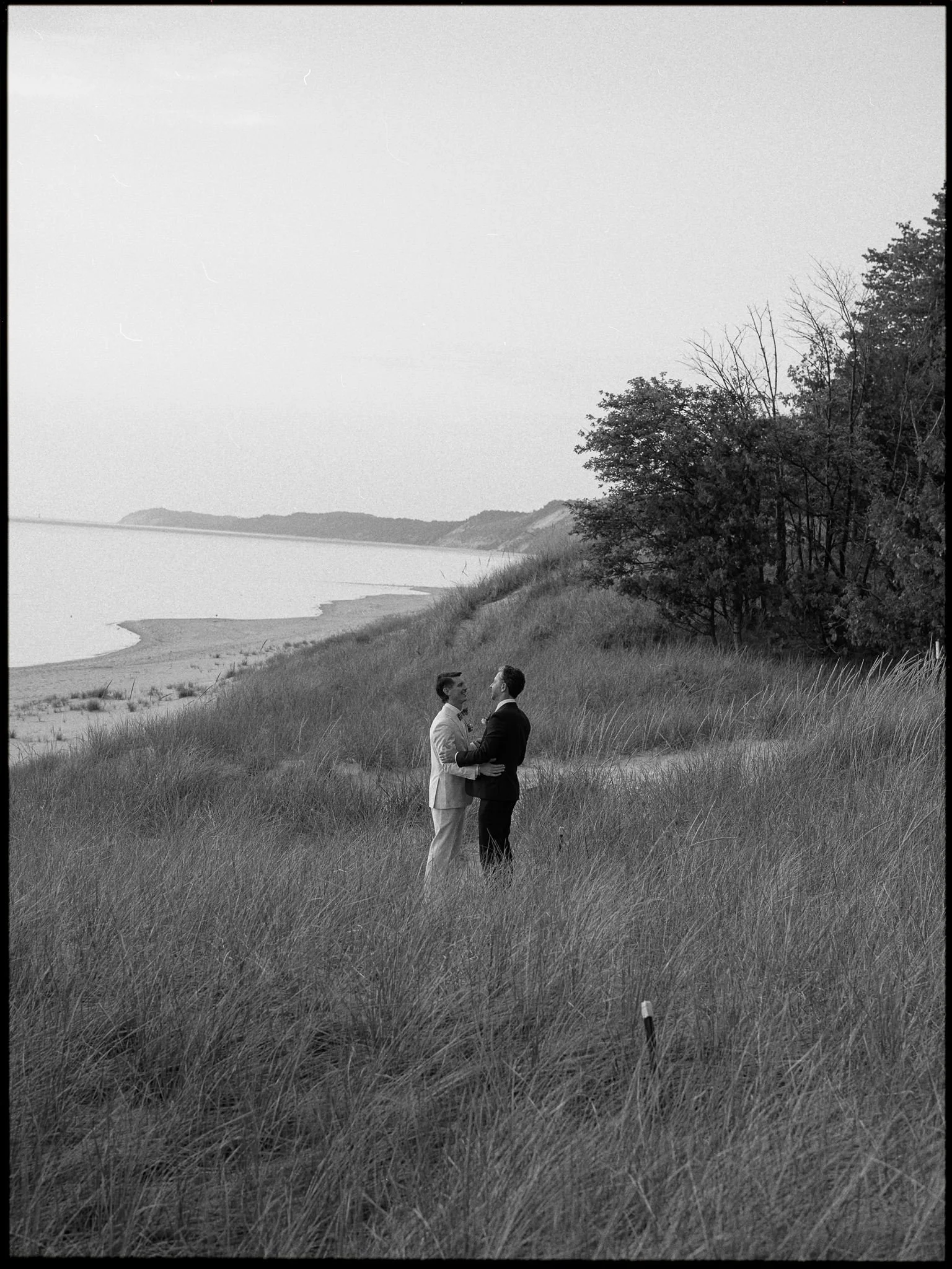 Two men in formal suits holding hands and facing each other in a grassy field by the water, with trees in the background, black and white photo.