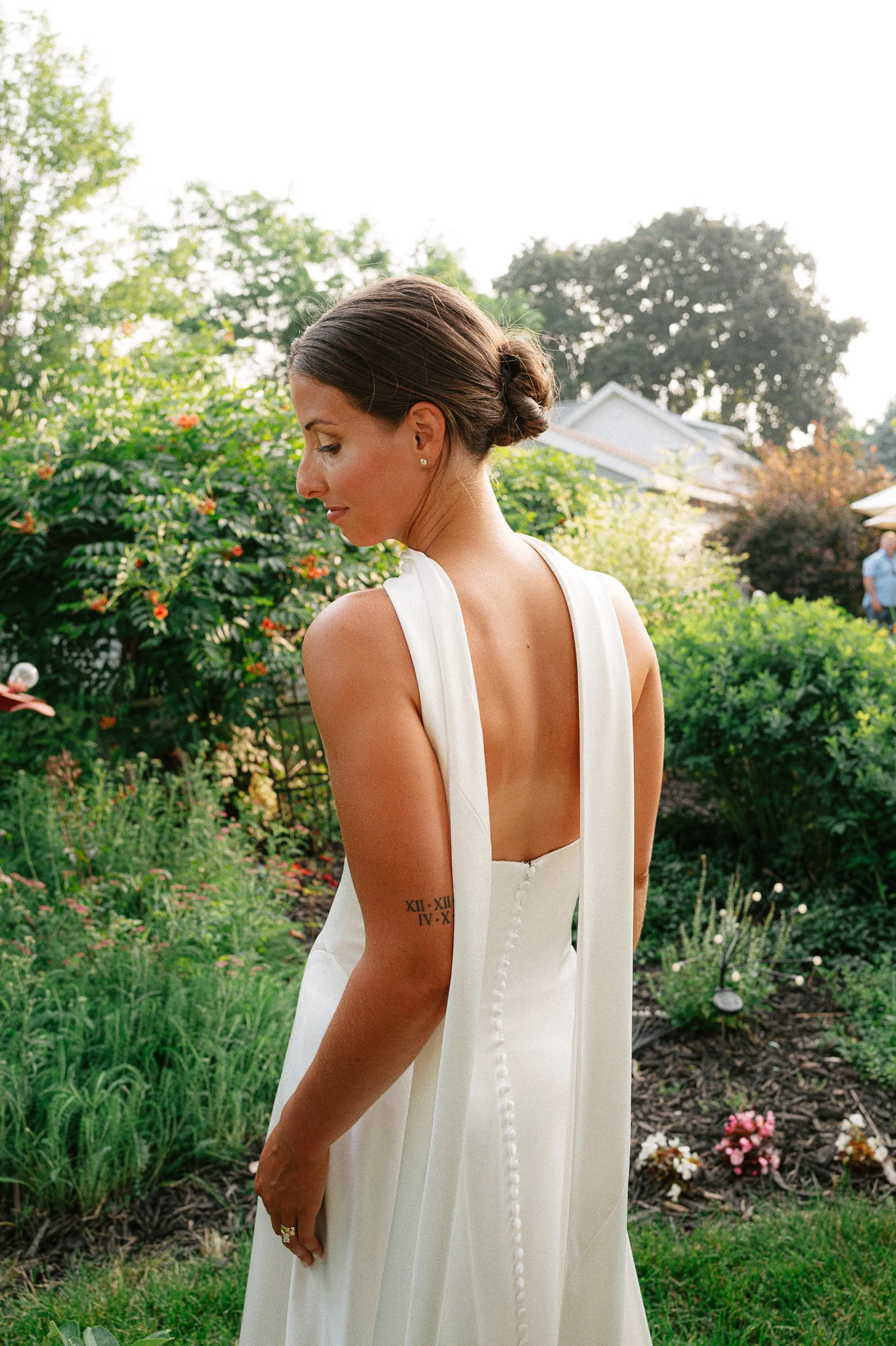 A bride with dark brown hair in a low bun, wearing a white wedding dress with a deep back and row of buttons, standing in a lush garden with greenery and flowers, looking down.