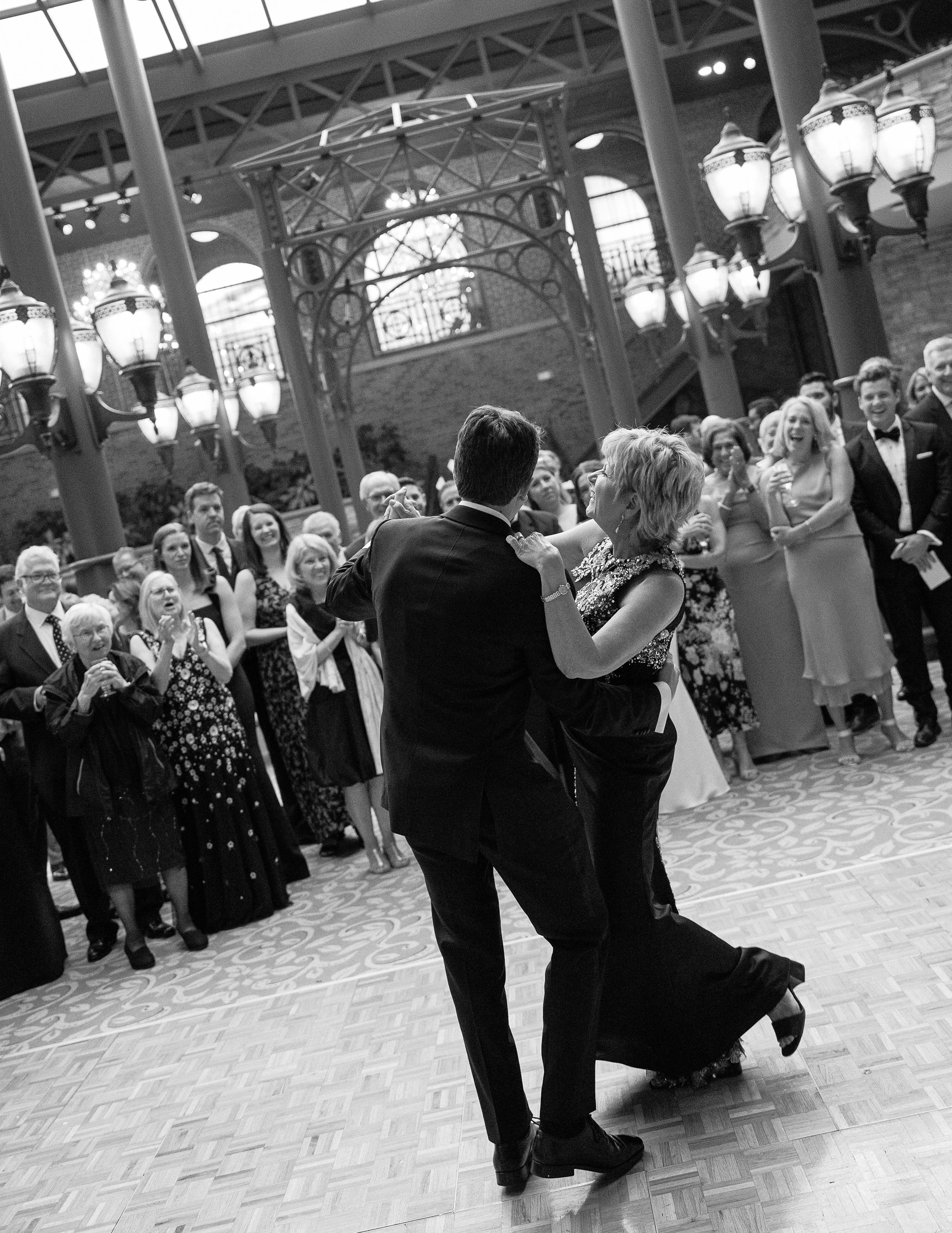 A black and white photo of a couple dancing in the center of a large, elegant hall surrounded by onlookers.