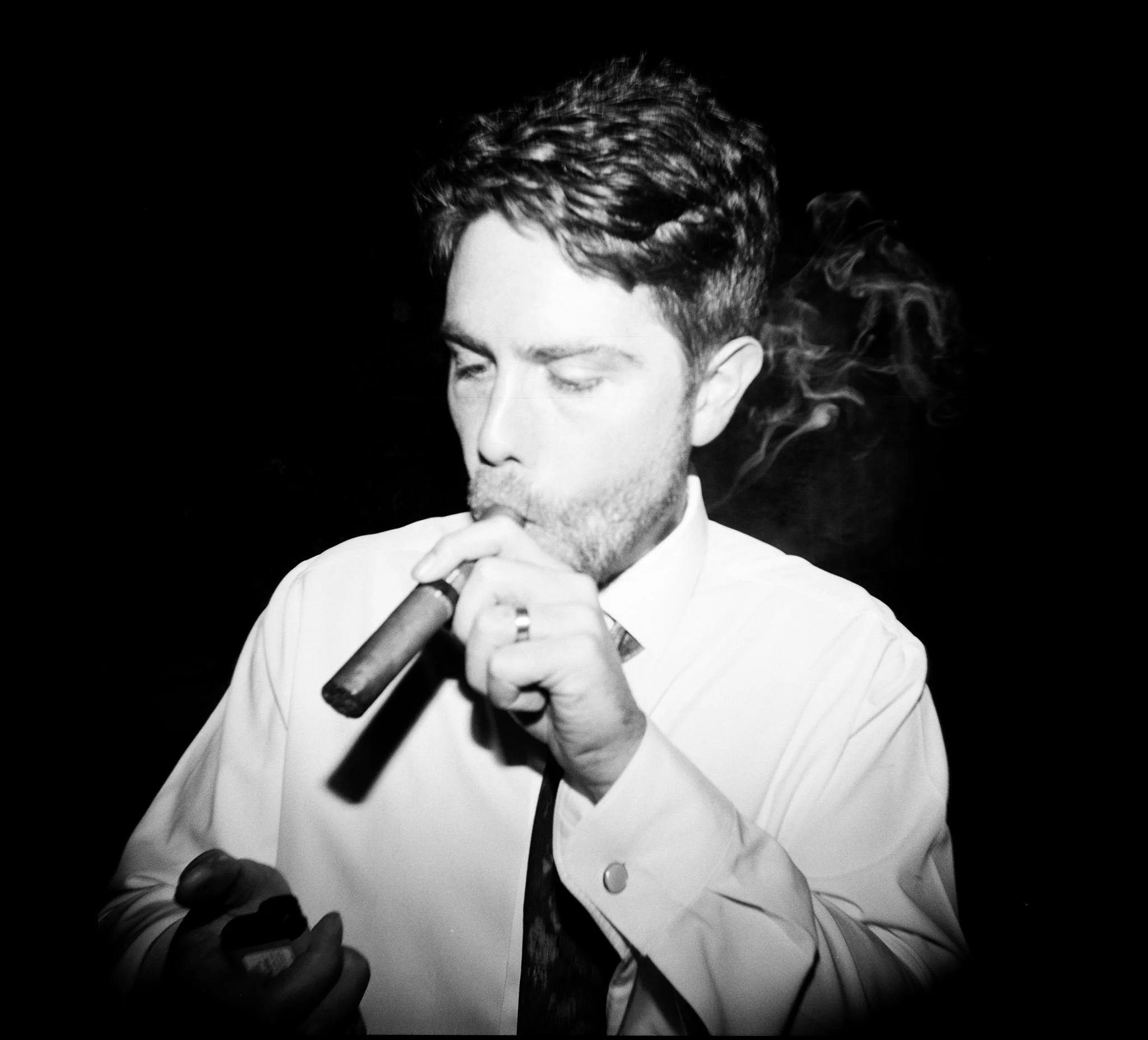 A black and white photo of a man with styled dark hair and a beard, wearing a white shirt, smoking a cigar, and looking at his phone over a dark background.