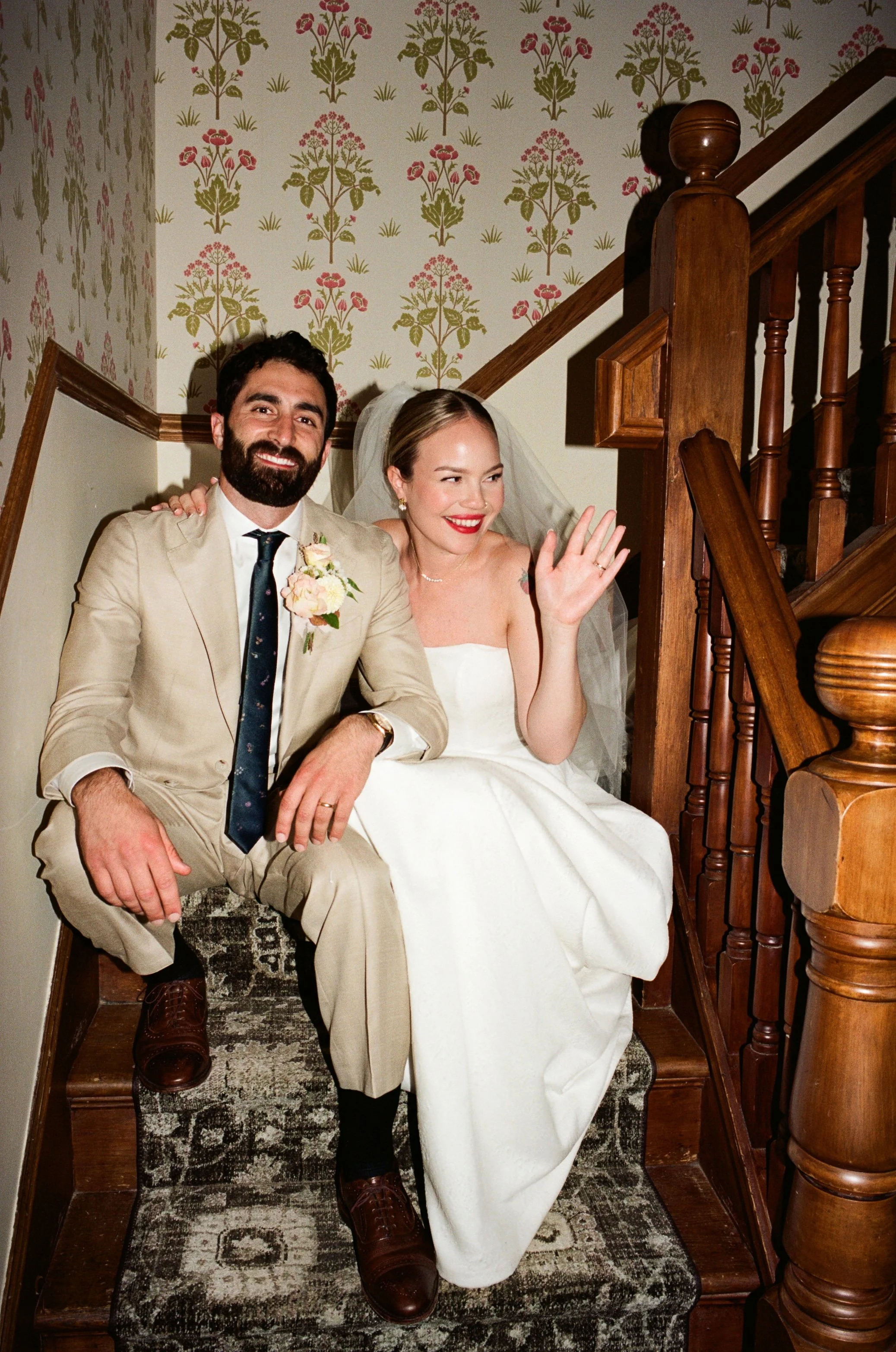 A bride and groom sitting on a staircase, smiling and waving. The bride is wearing a white strapless wedding dress and a veil, and the groom is wearing a beige suit with a dark tie and a boutonnière. The background features vintage floral wallpaper.
