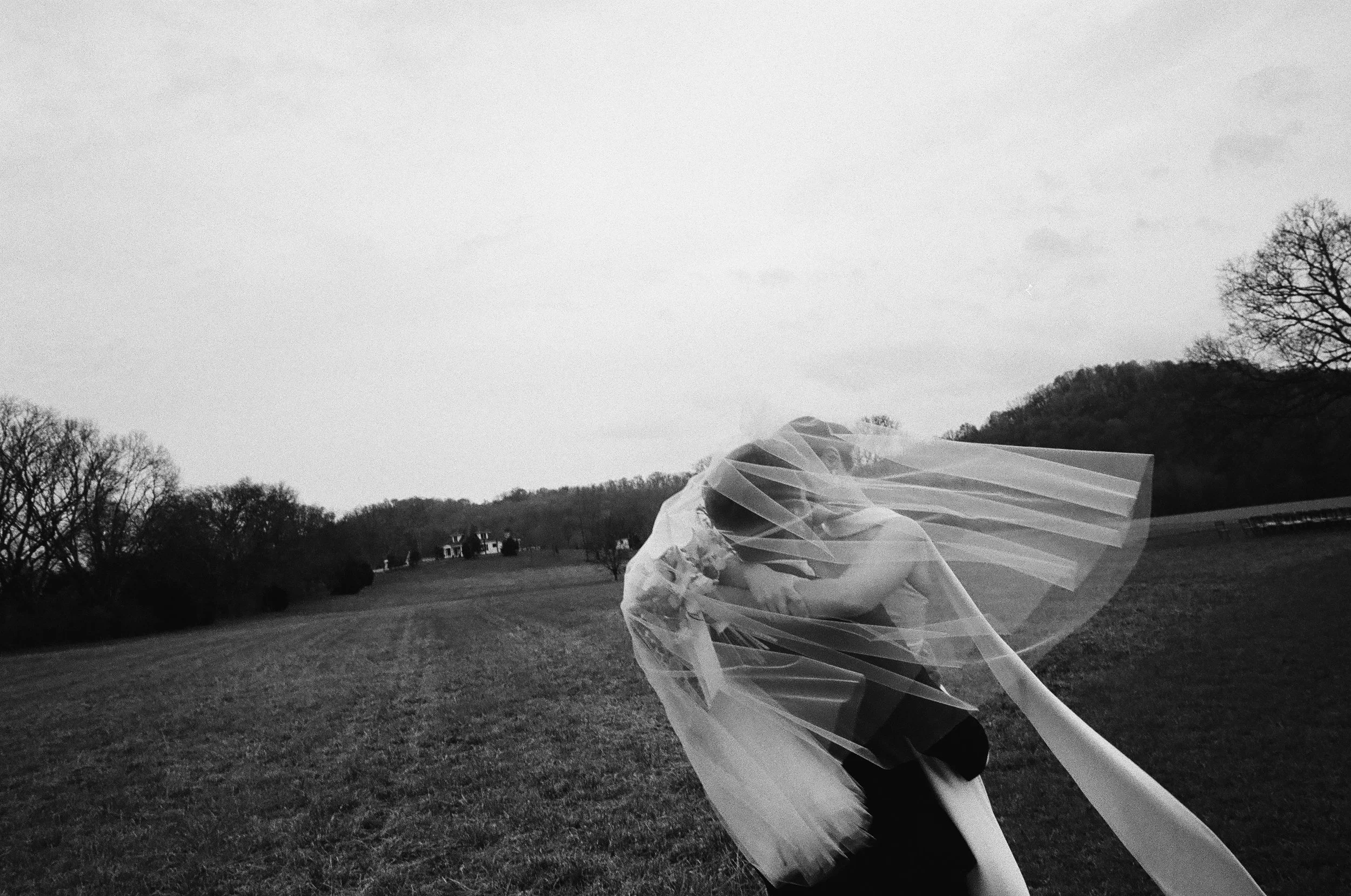 A person standing outdoors in a field with their face and body wrapped in a flowing, translucent fabric or veil, with trees and a hill in the background under a cloudy sky.