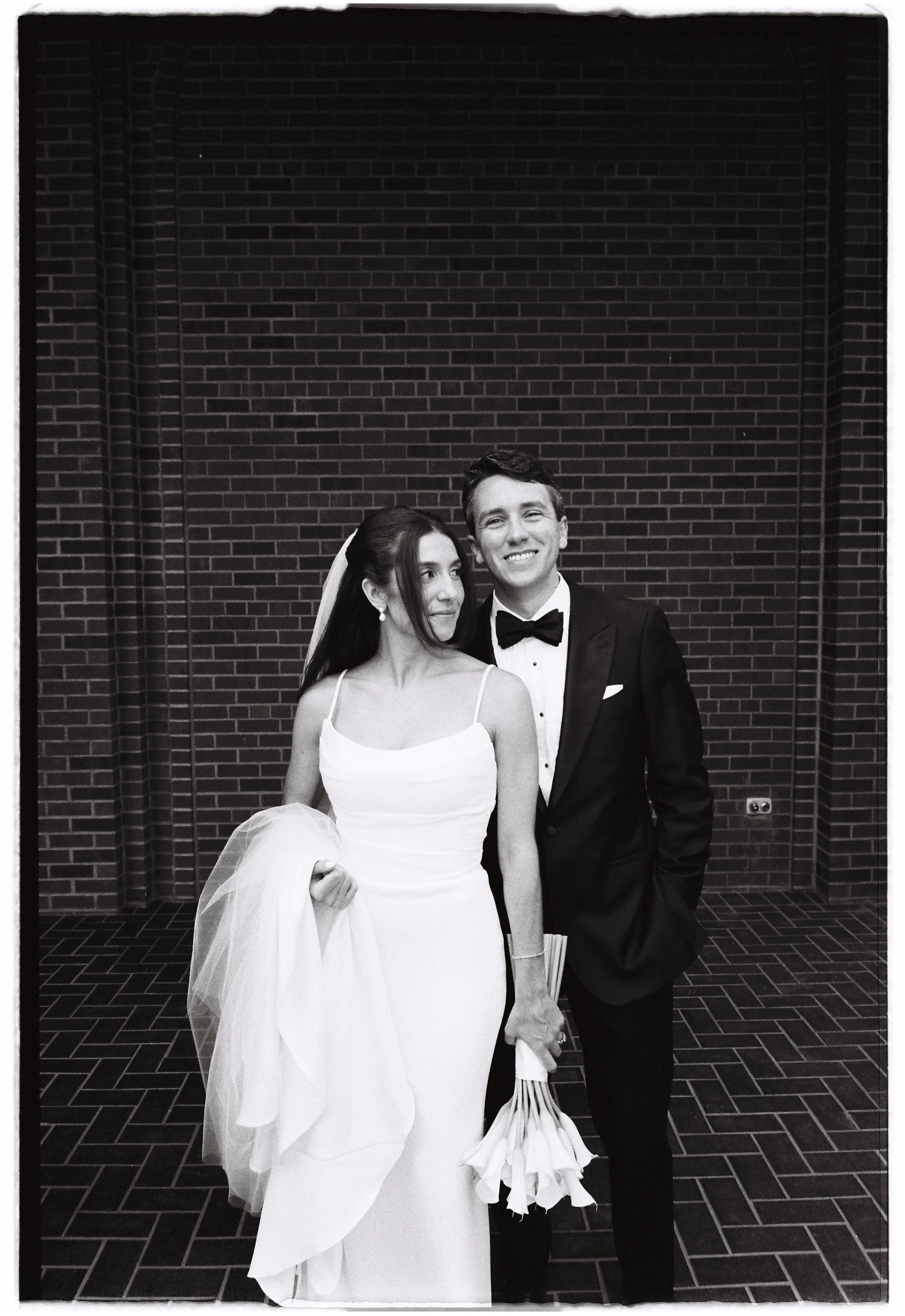 A bride and groom dressed in wedding attire, standing together, holding flowers, outdoors in front of a brick wall.