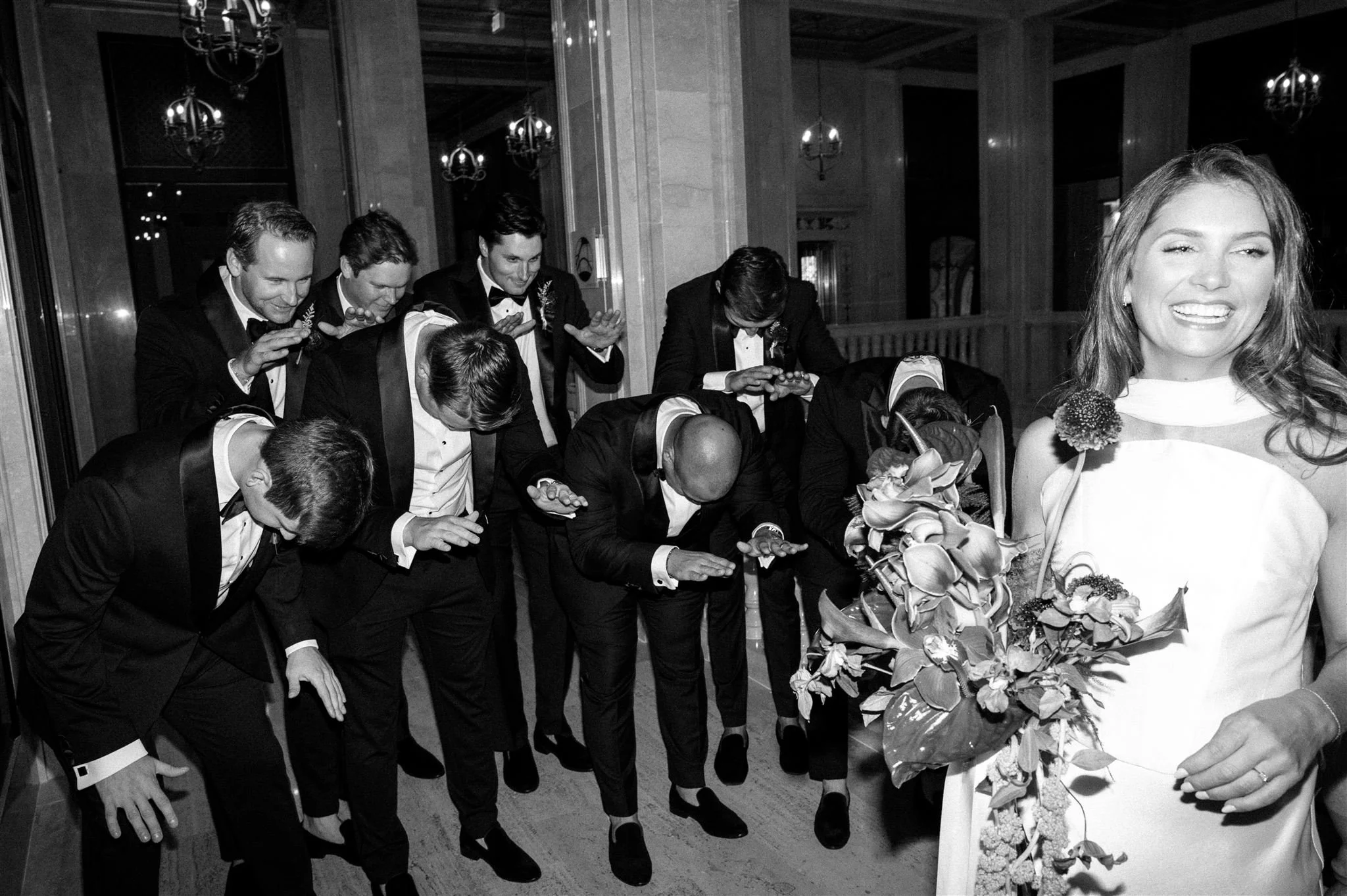 A woman holding a bouquet smiling at the camera, while a group of men dressed in tuxedos are bowing in the background.