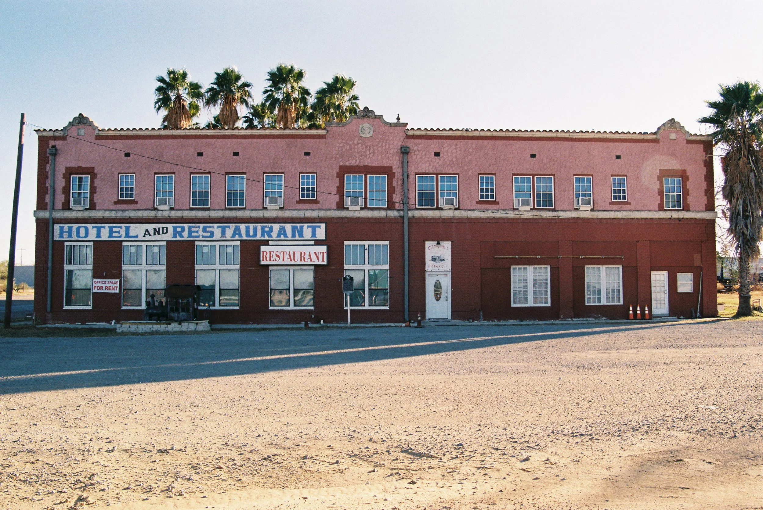 A two-story pink and red building with signs for a hotel and restaurant, located on a dirt lot with palm trees in the background.