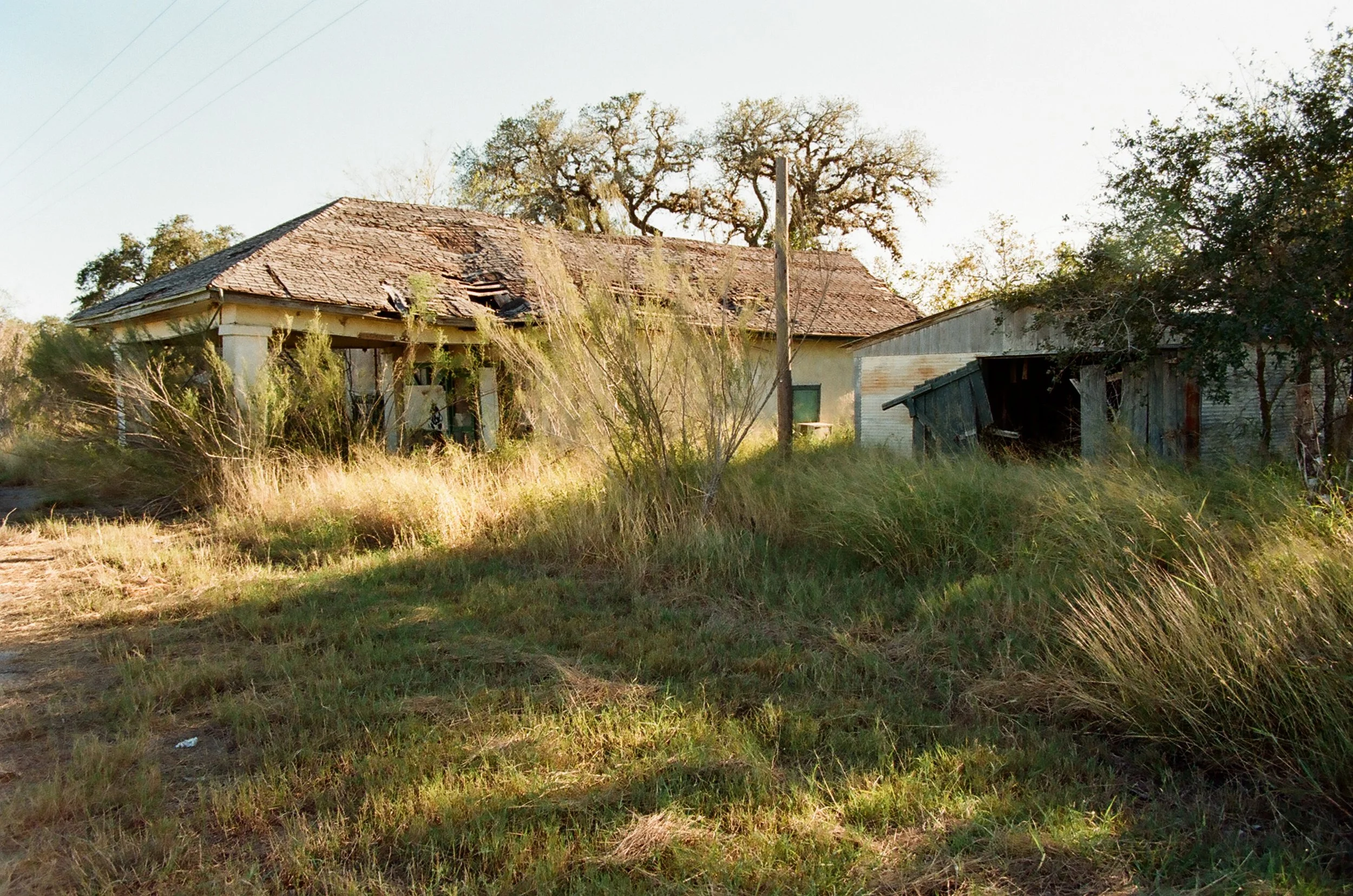 An abandoned house with a damaged roof, overgrown grass, and dilapidated structures in a rural setting.