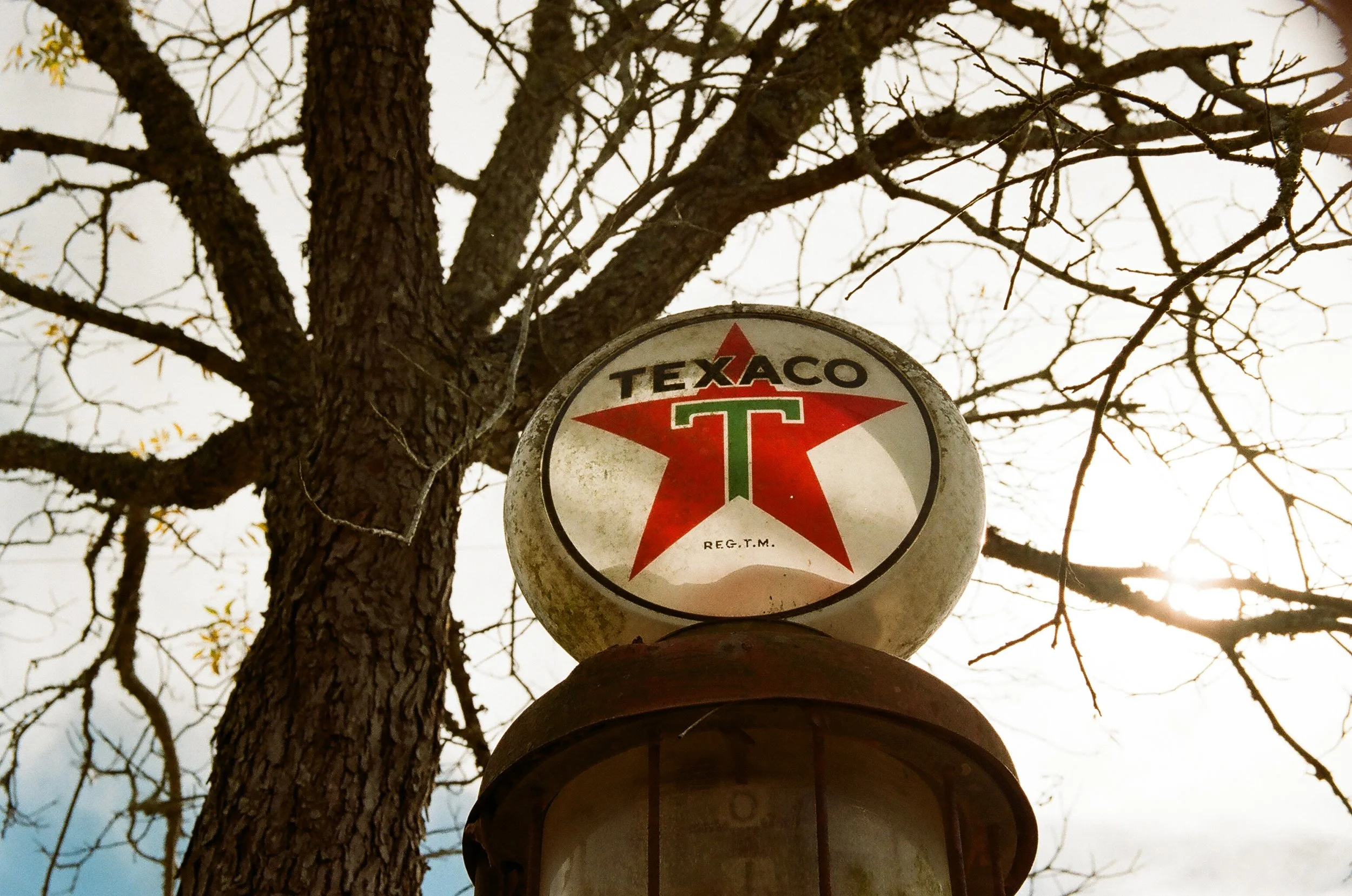 Vintage Texaco sign with red star logo and green T, mounted on a rusted pole against a background of leafless tree branches and a cloudy sky.