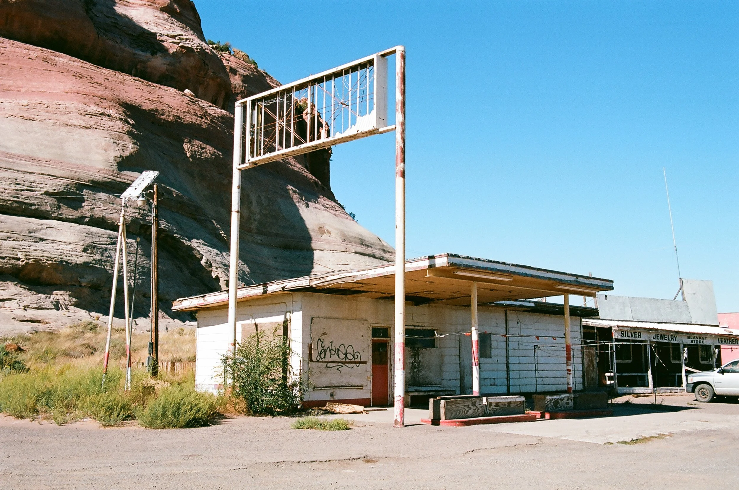 An abandoned small store with a white exterior, graffitied wall, boarded up windows, and a rusted sign. There is a parking lot in front, a white car, and a sign in the background reading 'Silver Jewelry, Blanket Store, Leather.' A rocky hill or mount