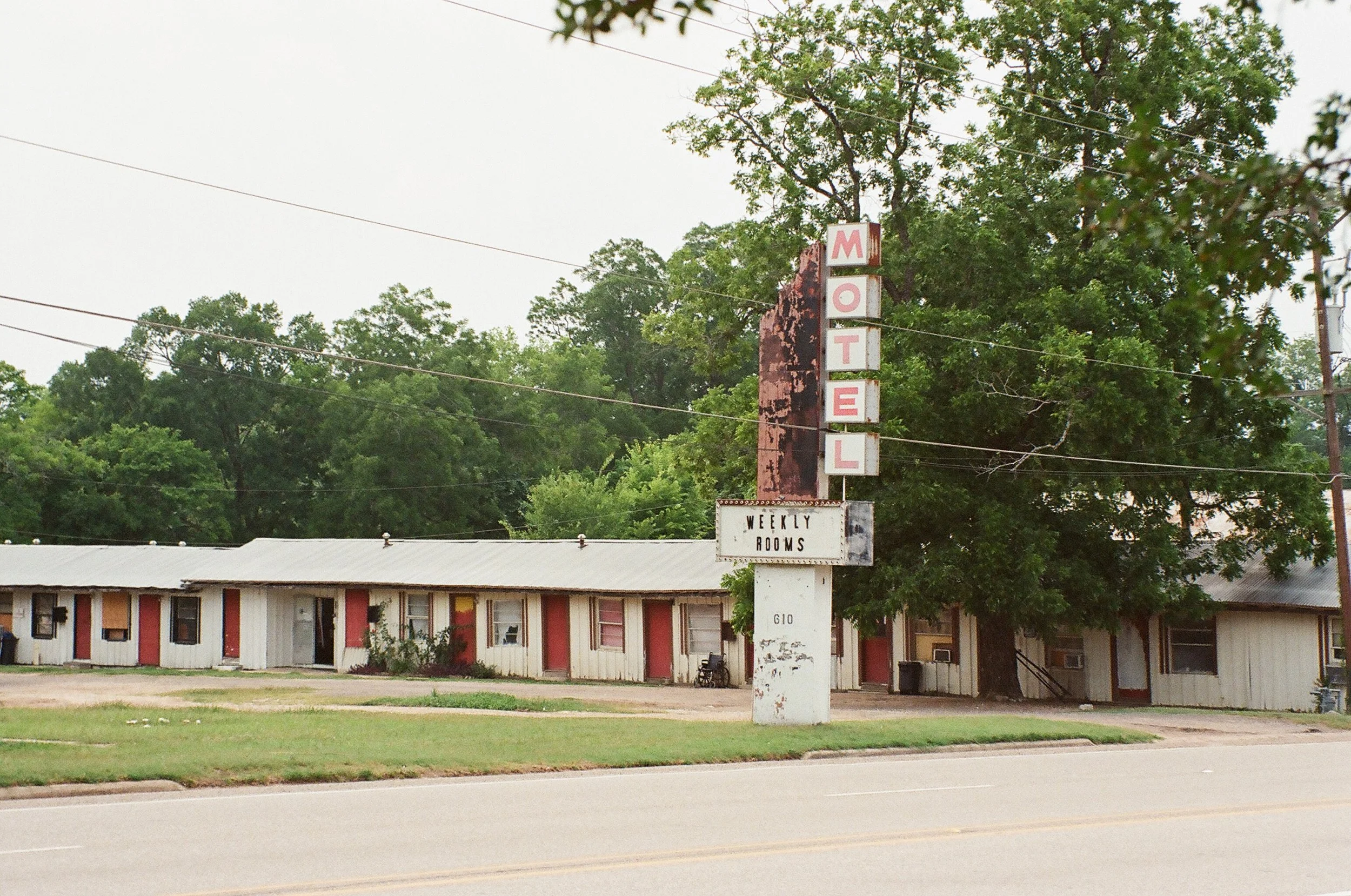 An old vintage motel sign with vertical letters spelling 'MOTEL' and a faded sign below reading 'Weekly Rooms', in front of a long, single-story motel building with a metal roof and mismatched doors, amid a backdrop of green trees.