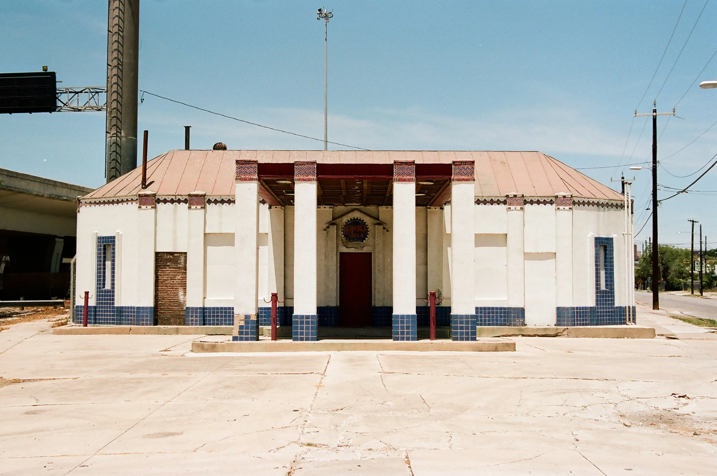 An old, small church building with a red metal roof and white walls, accented with blue tiles along the base and small side windows. The church has six large columns at the entrance and a small, arched doorway. The surrounding area is mostly paved wi
