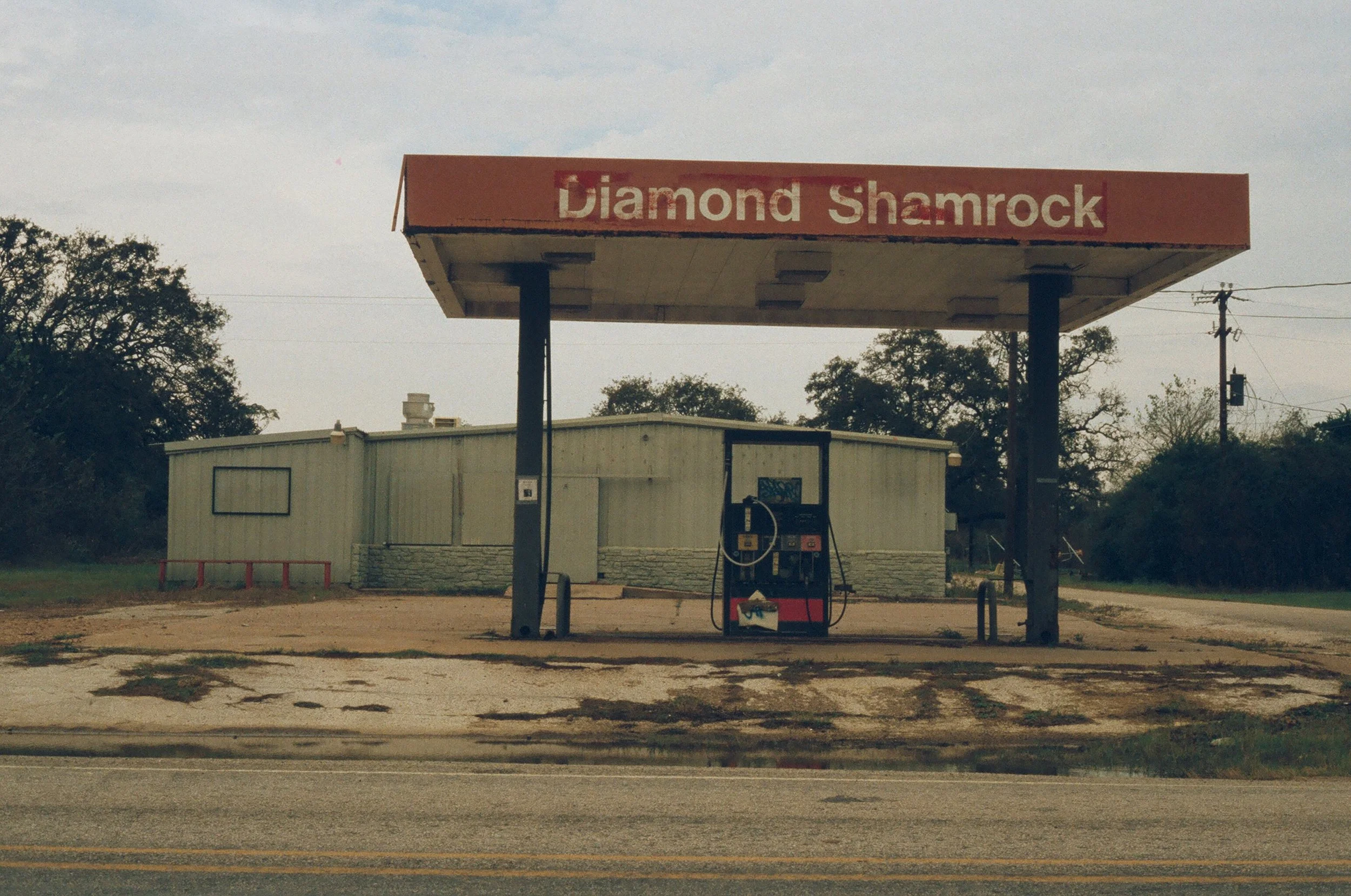 Abandoned gas station with a red canopy displaying the words 'Diamond Shamrock,' a single empty gas pump, and a small beige building in the background in a rural setting.