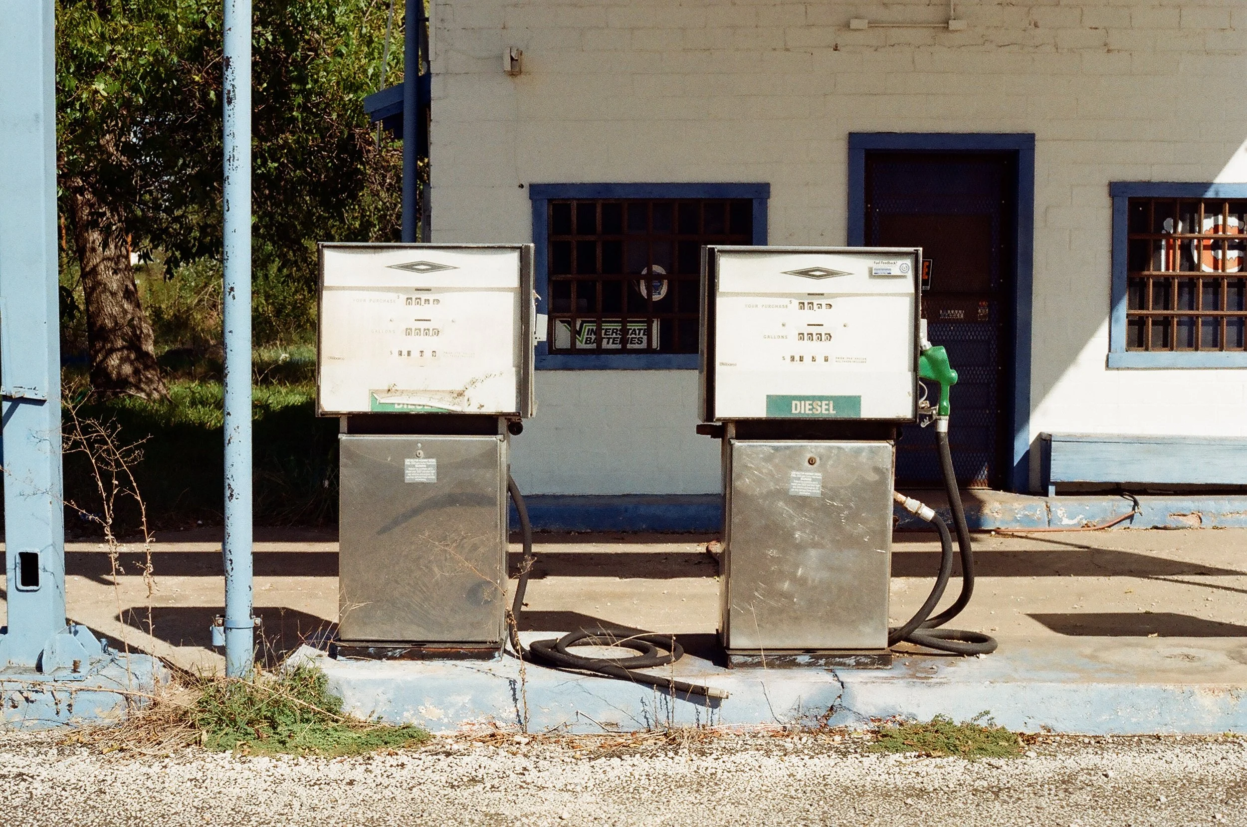 Two old gas pumps outside a small building with barred windows and a door. The building is painted white with blue trim. The area around the pumps is worn and overgrown with weeds.
