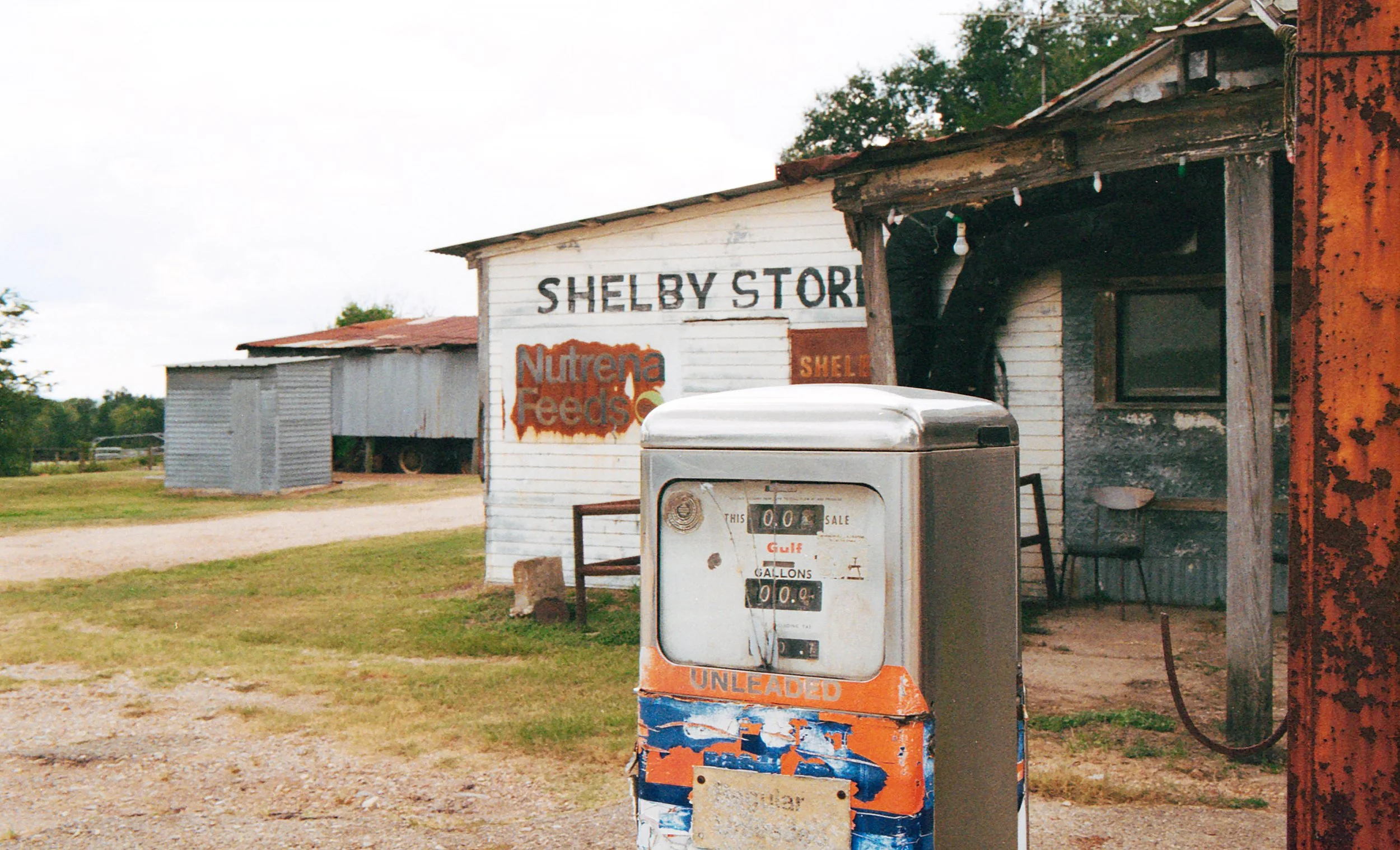 An old gas pump in front of a weathered building labeled 'Shelby Store', with rusted signboards including 'Nutrena Feed' and 'Shell'. The scene appears rural and nostalgic.