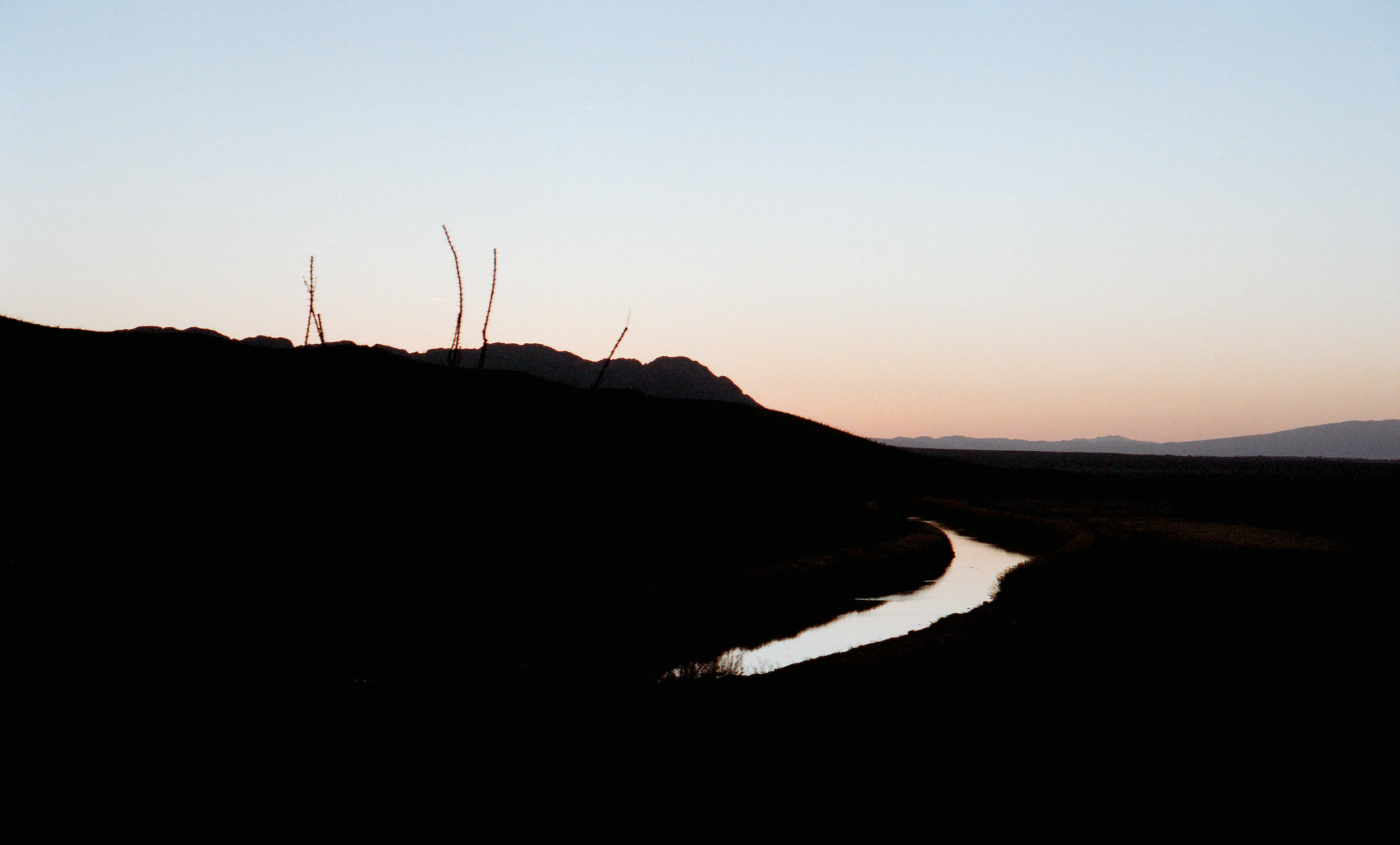 Silhouette of a winding river with hills in the background during sunset or sunrise, with a clear sky.