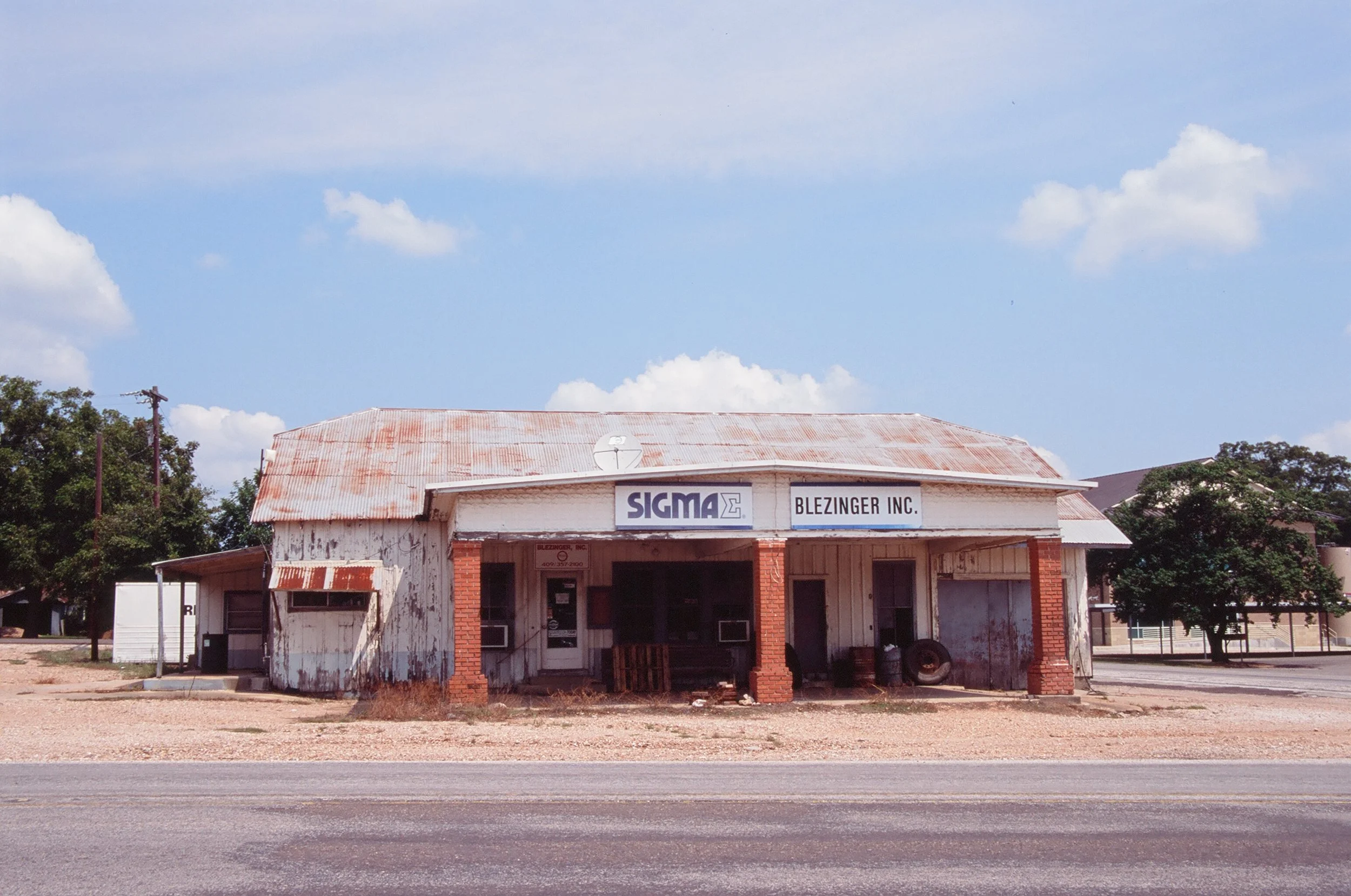 An old, weathered building with a rusted metal roof, white wooden siding, and brick columns, housing an unused business named 'Sigma Blezinger Inc.' on a mostly deserted street under a blue sky with scattered clouds.